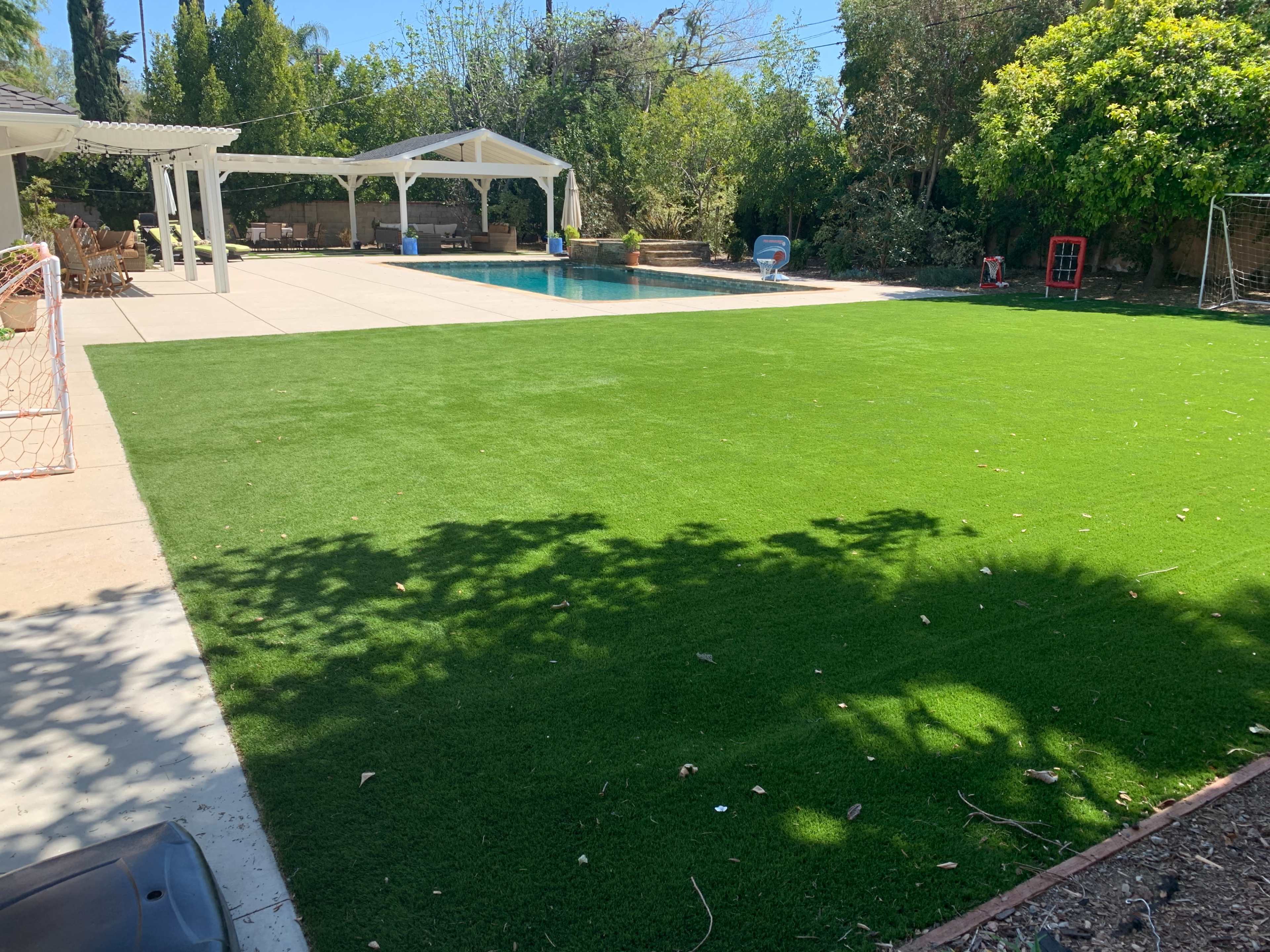 A well-maintained backyard features a swimming pool surrounded by artificial grass and shaded by trees.