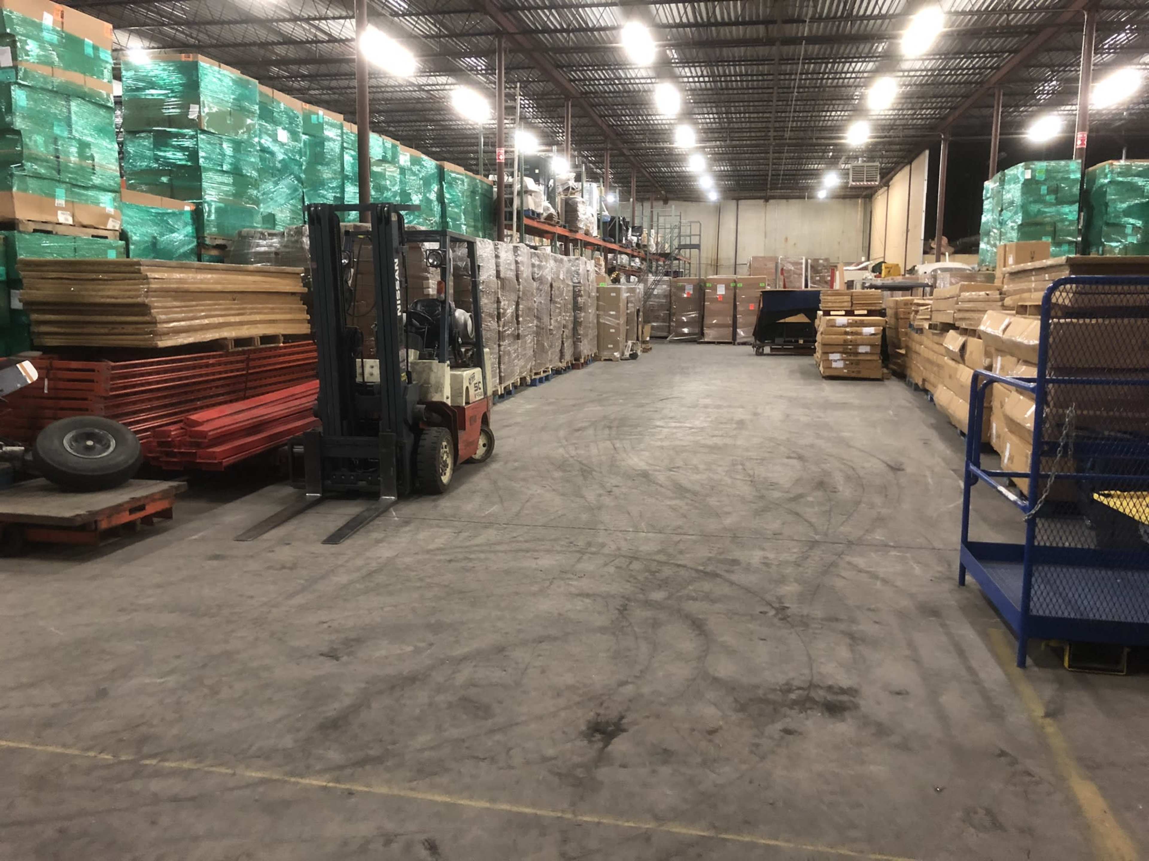 A warehouse interior with stacked pallets of goods on either side of a wide, empty central aisle and a forklift positioned on the left.
