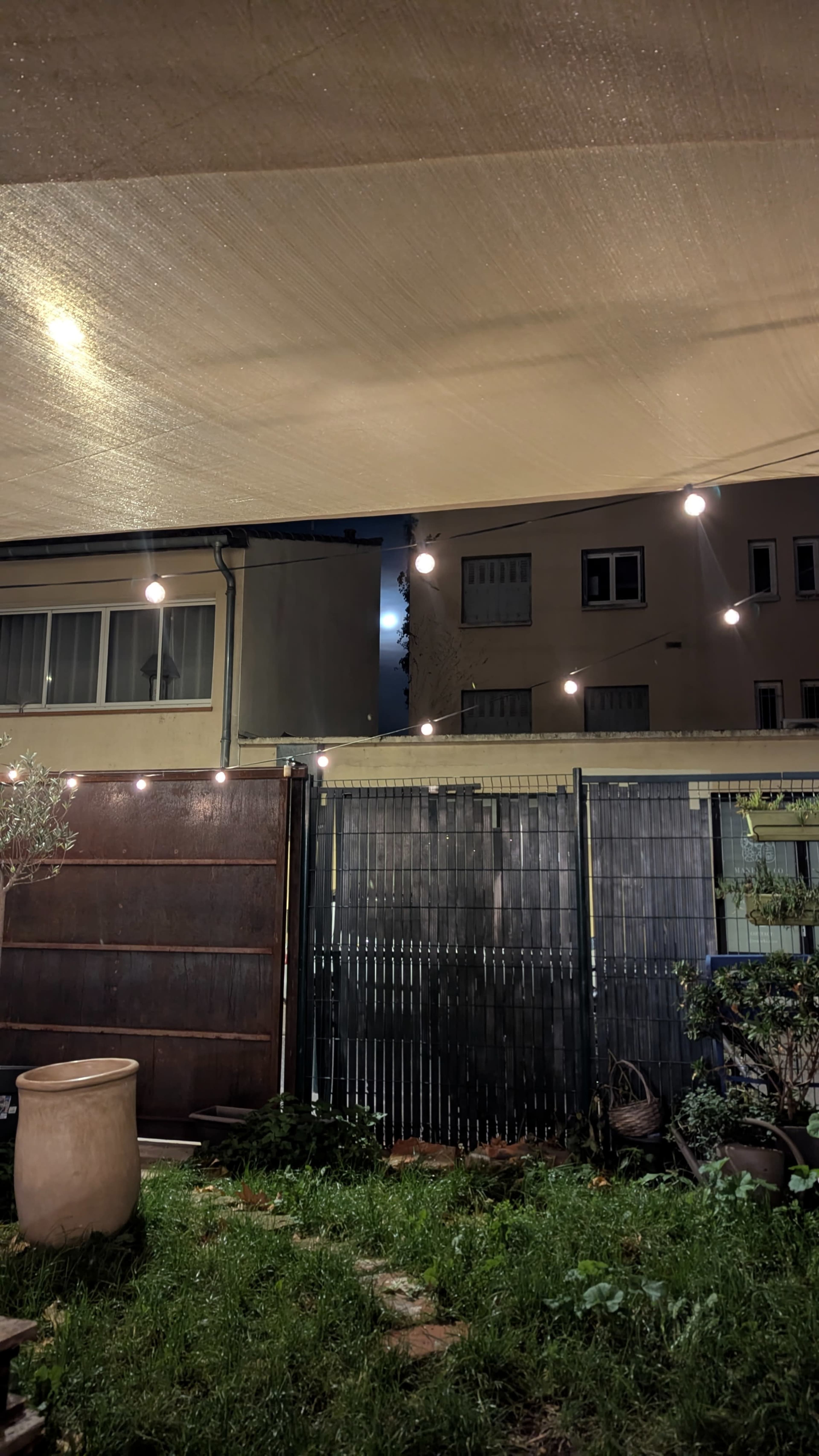 The image shows a garden area with a gravel ground, illuminated by string lights, a large planter, and a backdrop of buildings partially obscured by a canopy.
