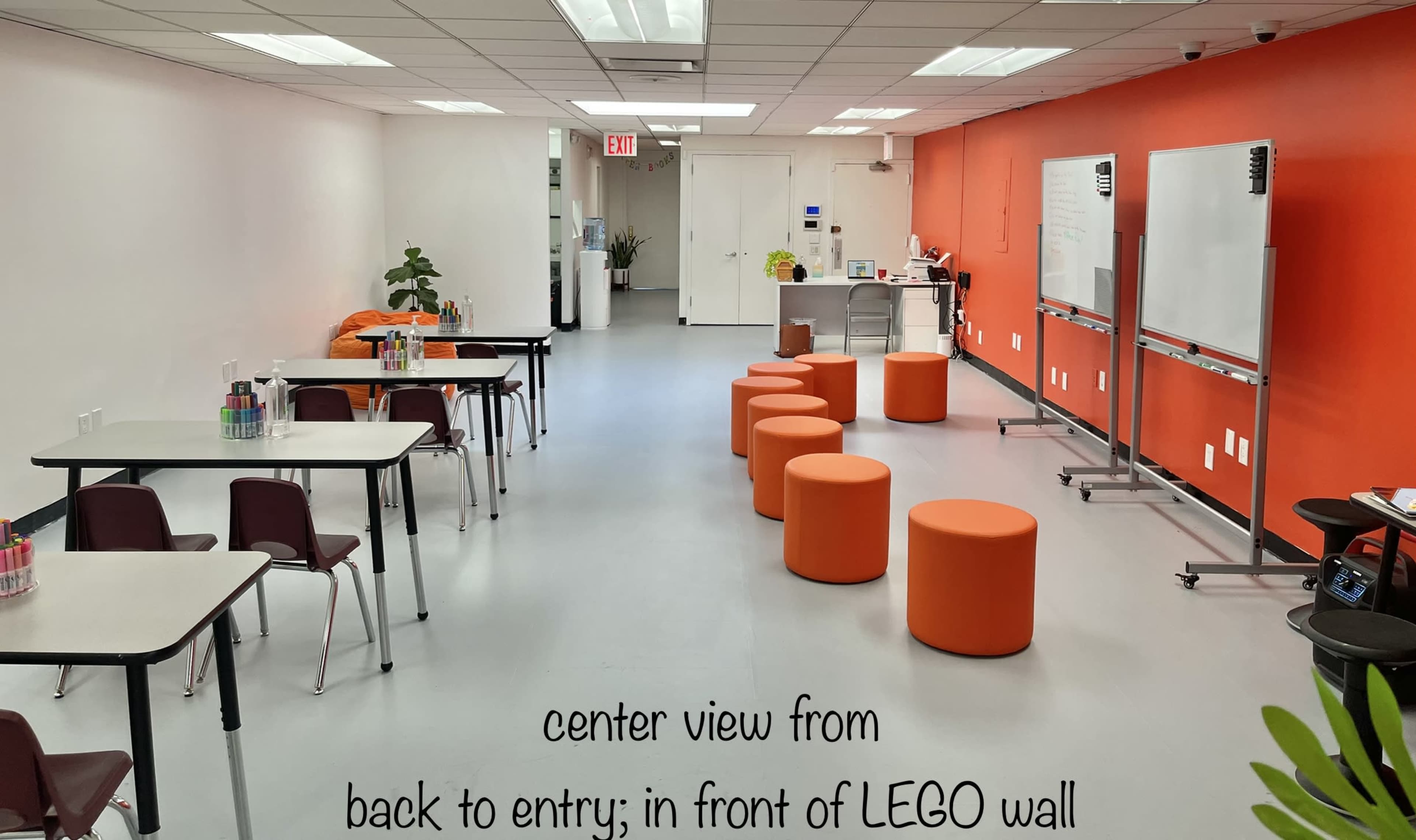 A bright room with orange walls, featuring tables and chairs on one side and orange stools arranged in the center, while a LEGO wall and whiteboards are visible on the other side.