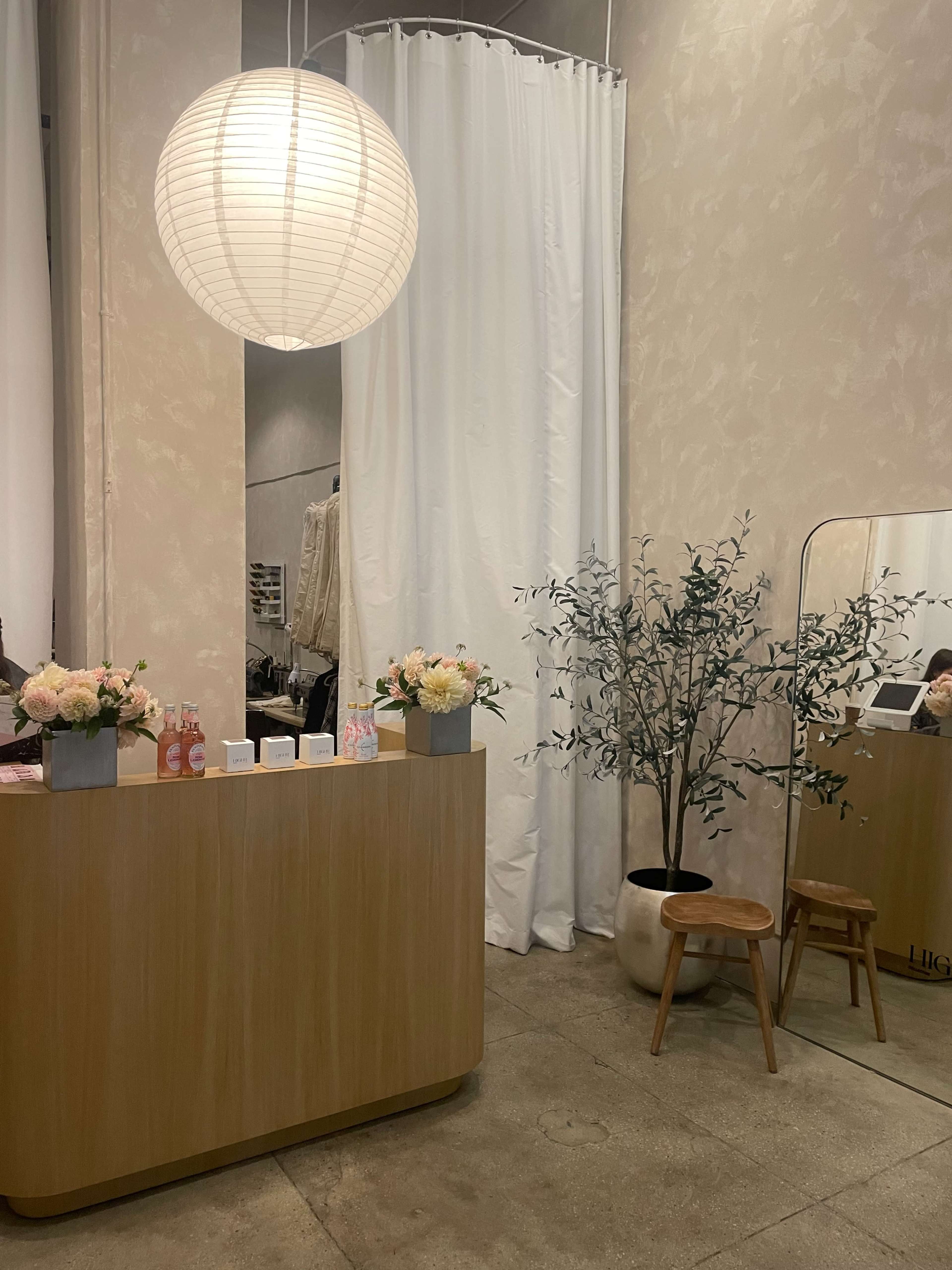 A bright interior of a retail space featuring a wooden reception desk, a large round pendant light, a mirror, a potted plant, and a white curtain dividing the area.