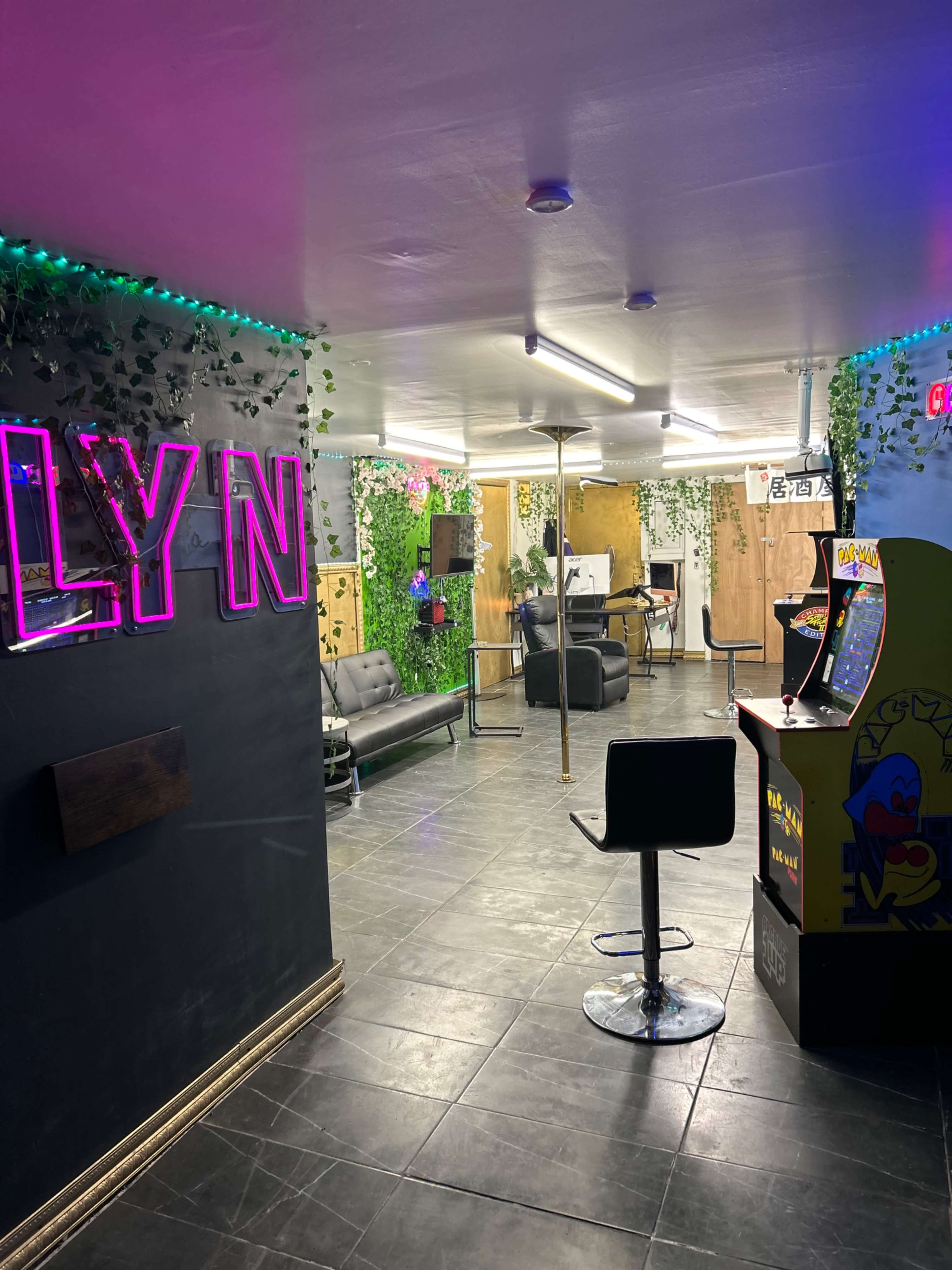 A modern interior space with neon signs, a barbershop chair, a futon, and an arcade machine, all surrounded by greenery and colorful lighting.