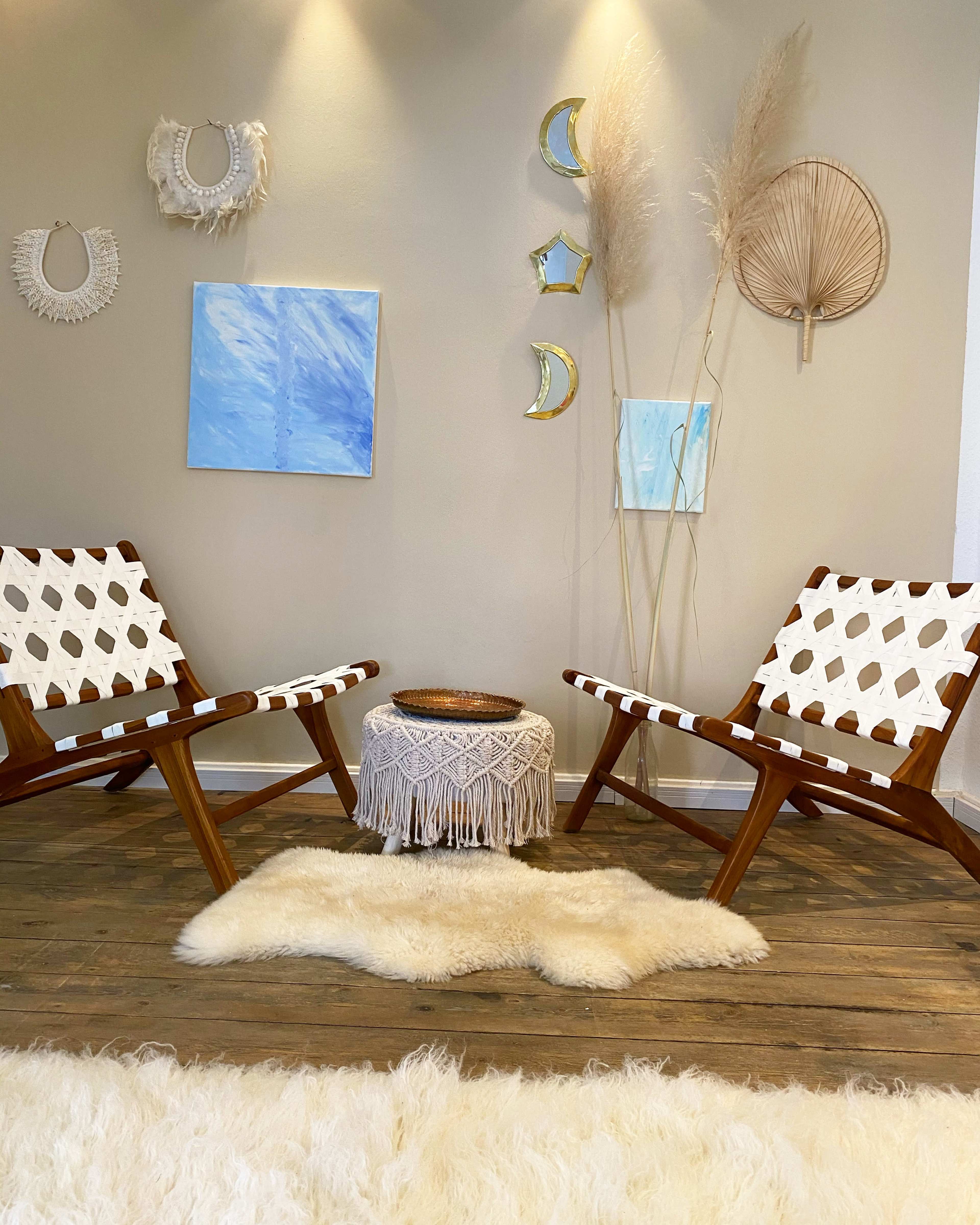 The image shows a cozy room with two wooden chairs, a small round table with a decorative cover, and wall art that includes paintings and woven decorations.