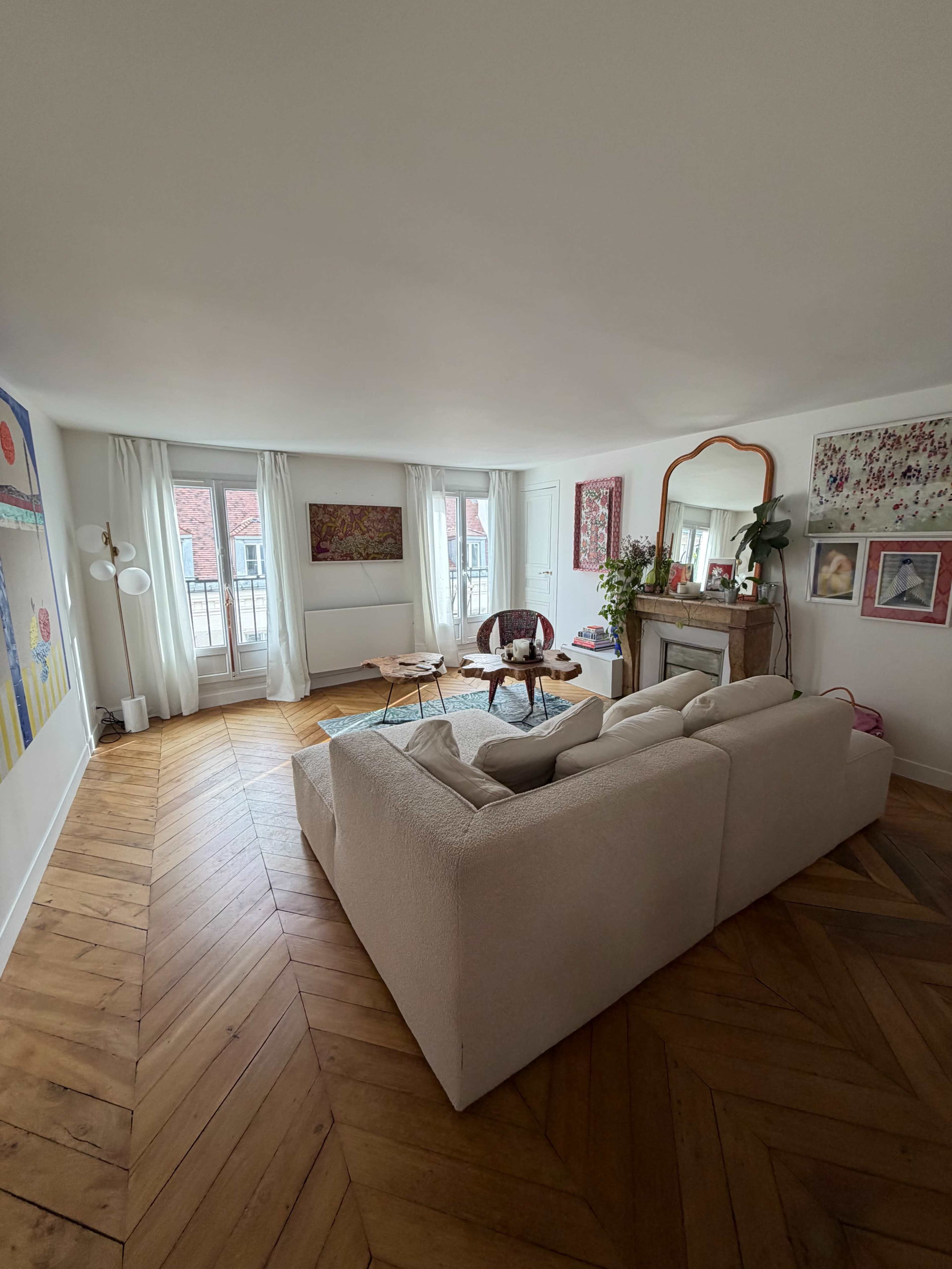 The image shows a well-lit living room with a beige couch, two wooden coffee tables, a decorative mirror, and large windows covered with white curtains.
