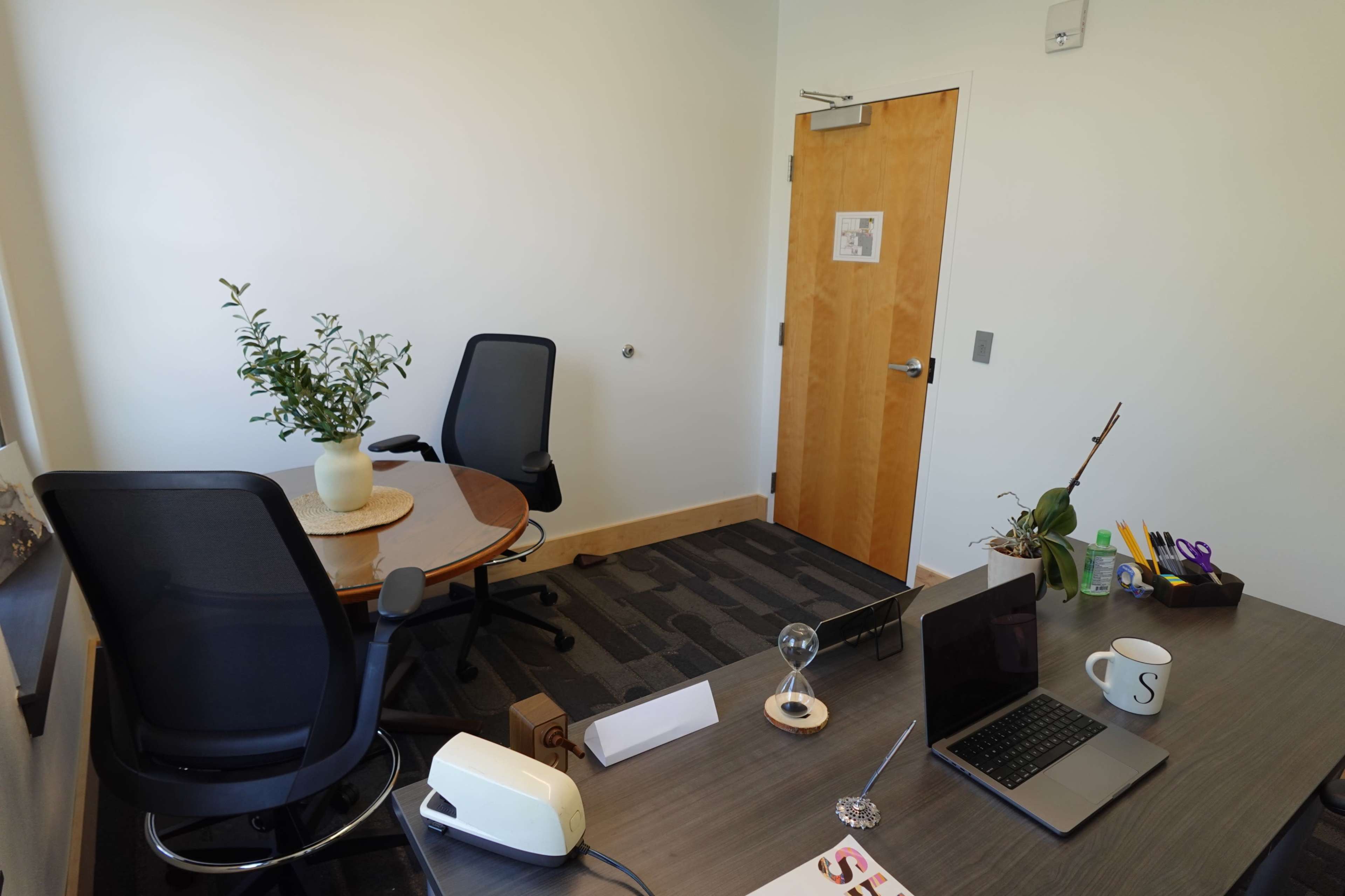 The image shows a neatly organized office space with two chairs, a round table, a desk with a laptop, a mug, and a small plant.