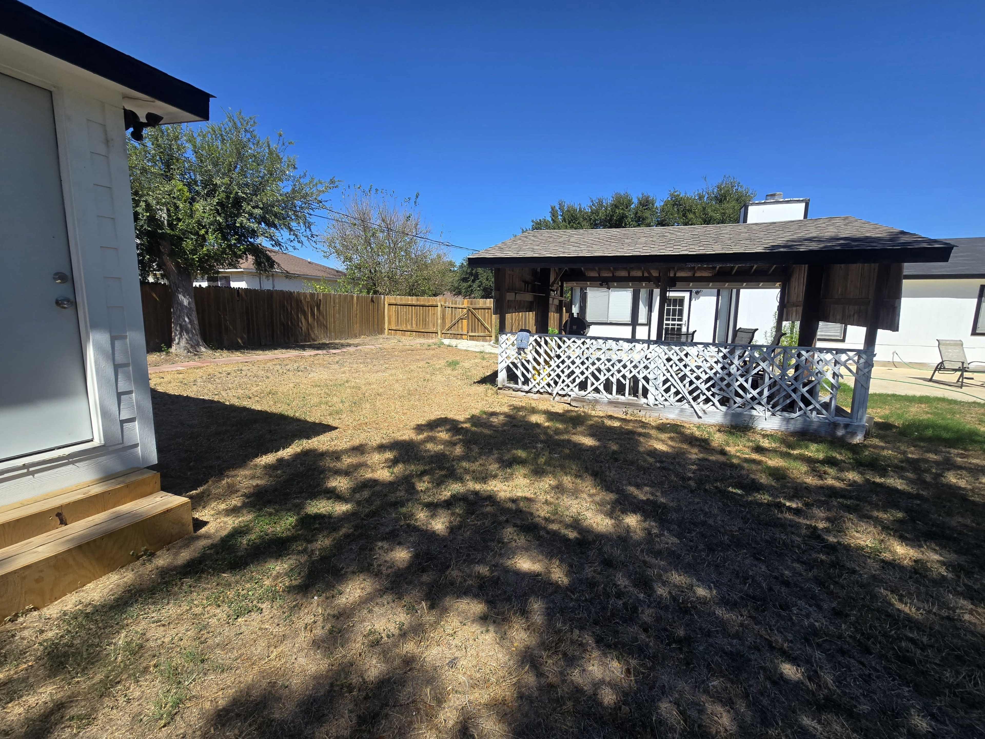 The image shows a backyard with a gazebo, a small building on the left, and a wooden fence in the background under a clear blue sky.