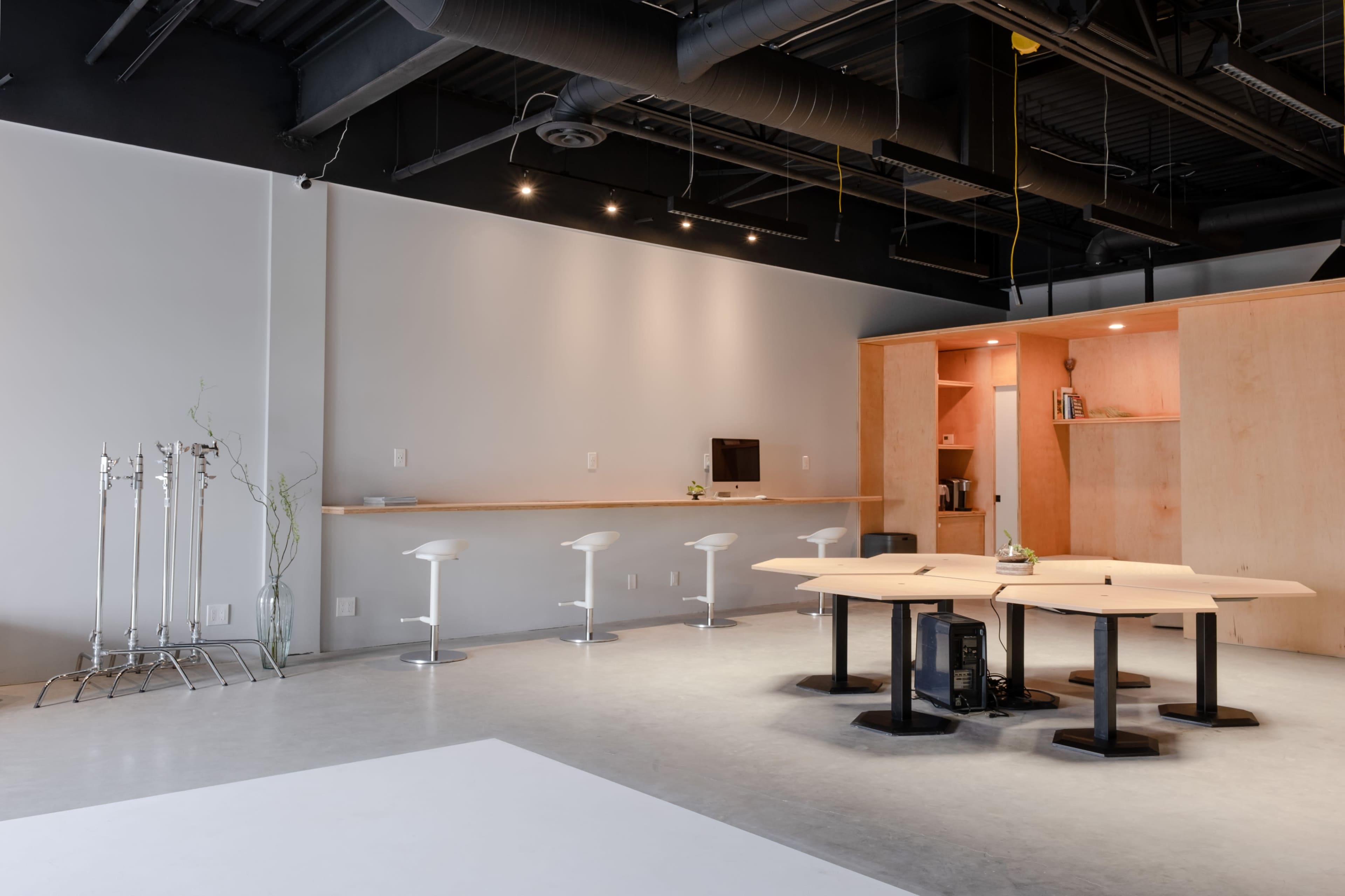 A modern open office space featuring a wooden meeting table, white bar stools, and minimalistic cabinetry against a light gray wall.
