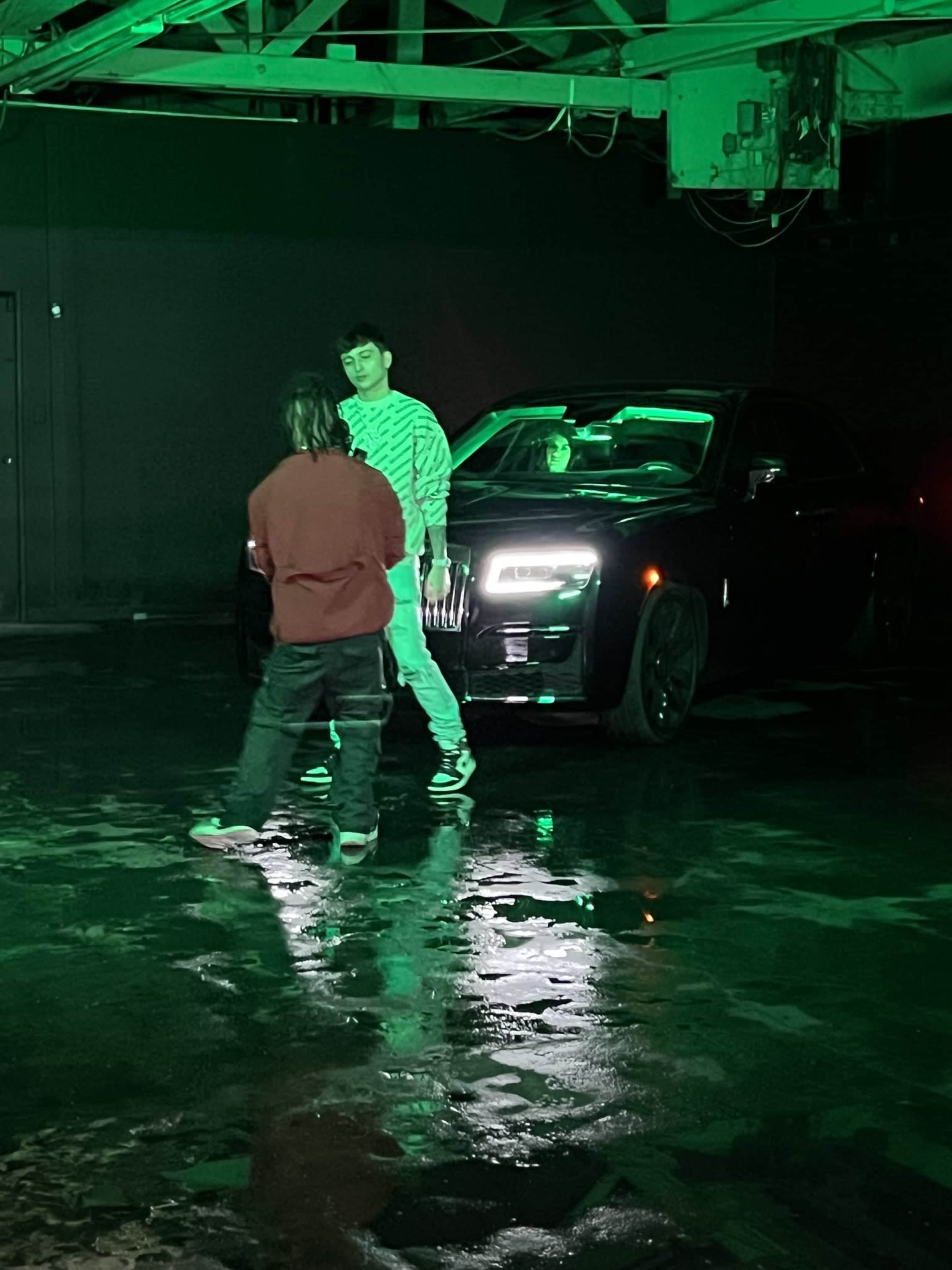 Two individuals stand in front of a black car with green lighting in an industrial space.
