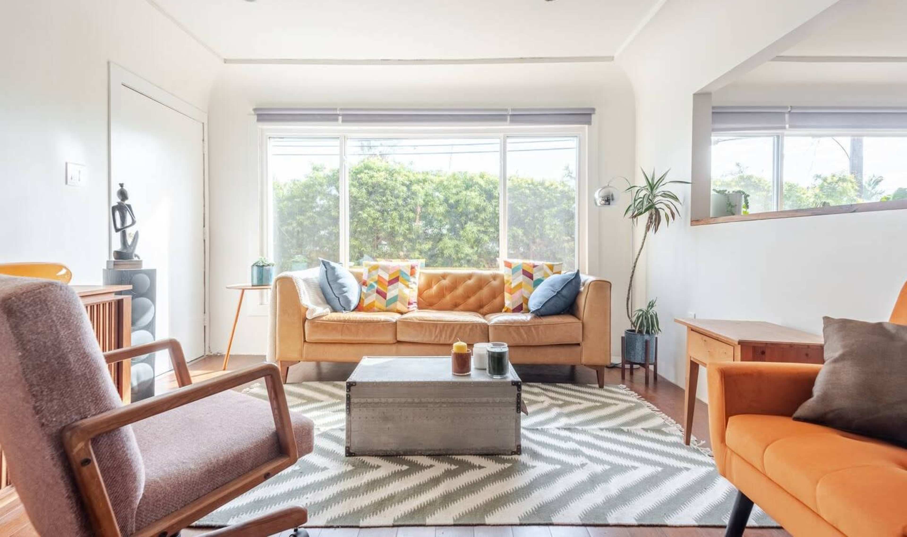 The scene depicts a modern living room with a brown couch, patterned cushions, a coffee table, and large windows letting in natural light.