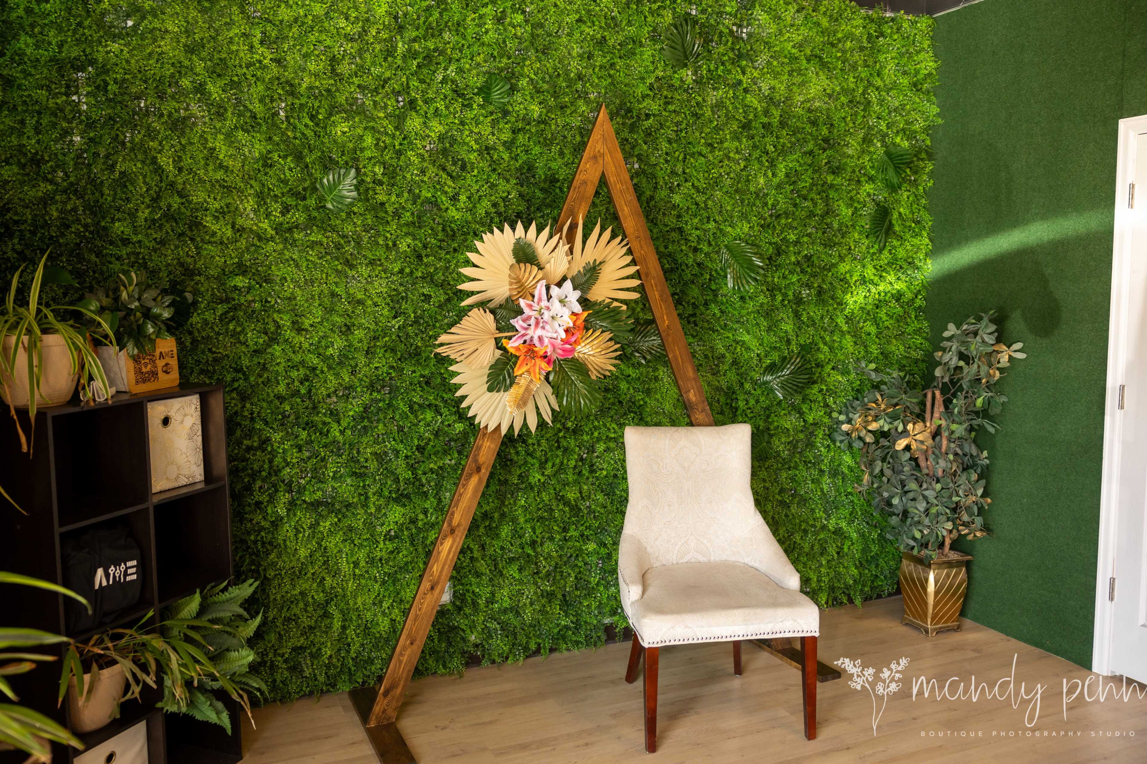 A wooden frame in the shape of an "A" stands against a green moss-covered wall, with a decorative floral arrangement and a white chair positioned nearby.