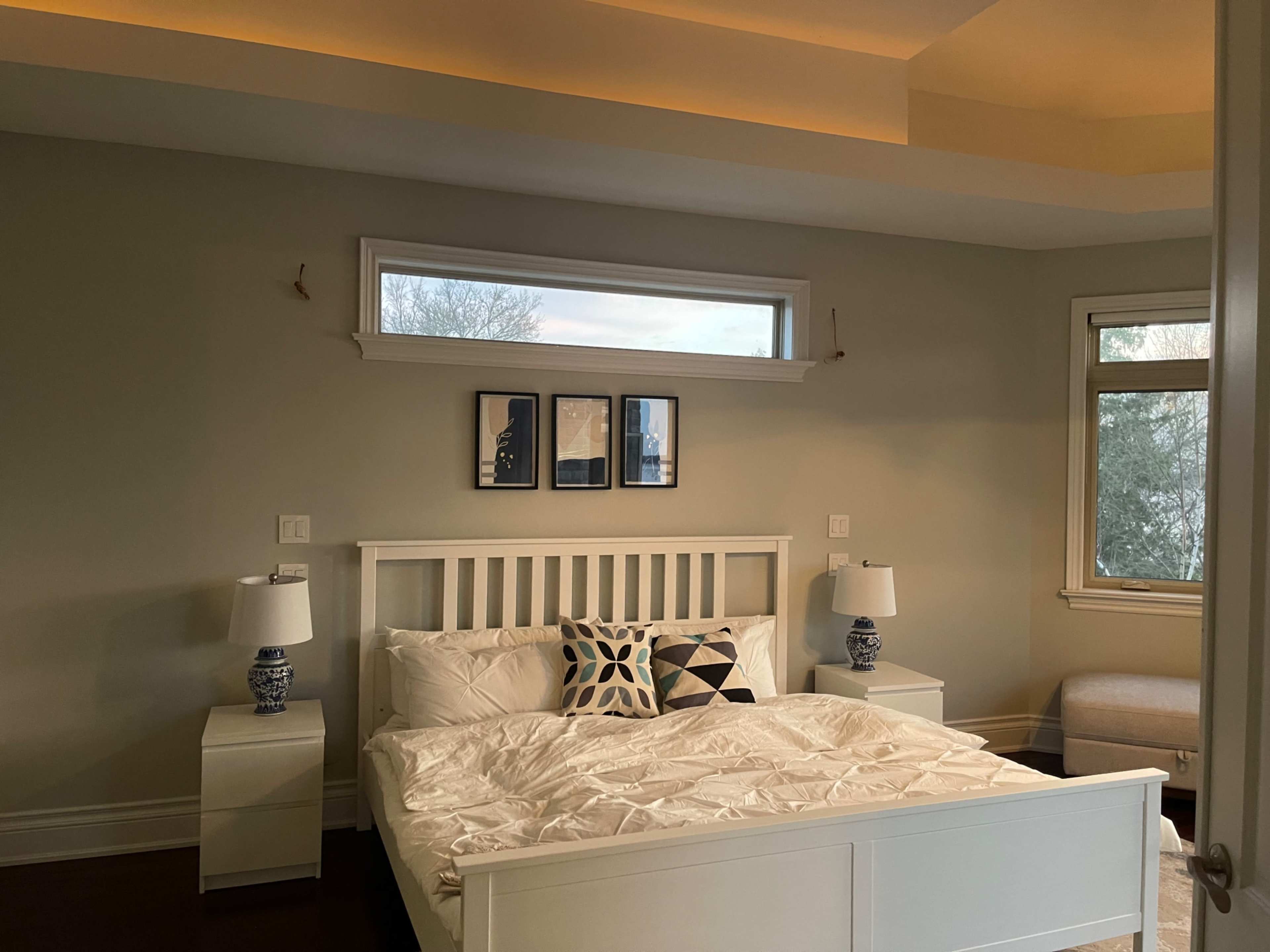 A neatly made bed is positioned between two bedside tables with lamps in a softly lit bedroom featuring large windows.