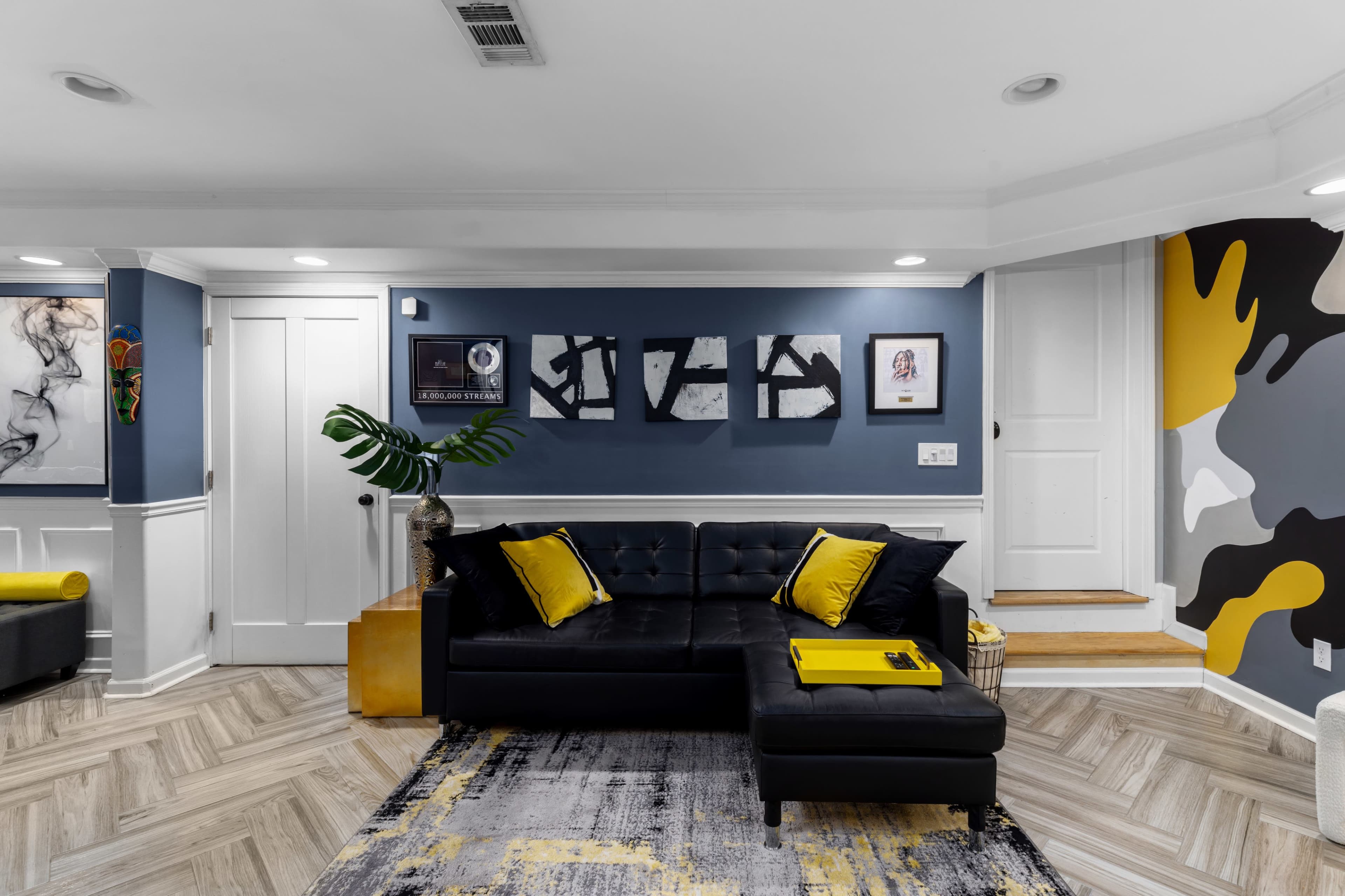 The image shows a modern living room featuring a black sectional sofa with yellow accents, wall art, and a patterned area rug on herringbone wood flooring.