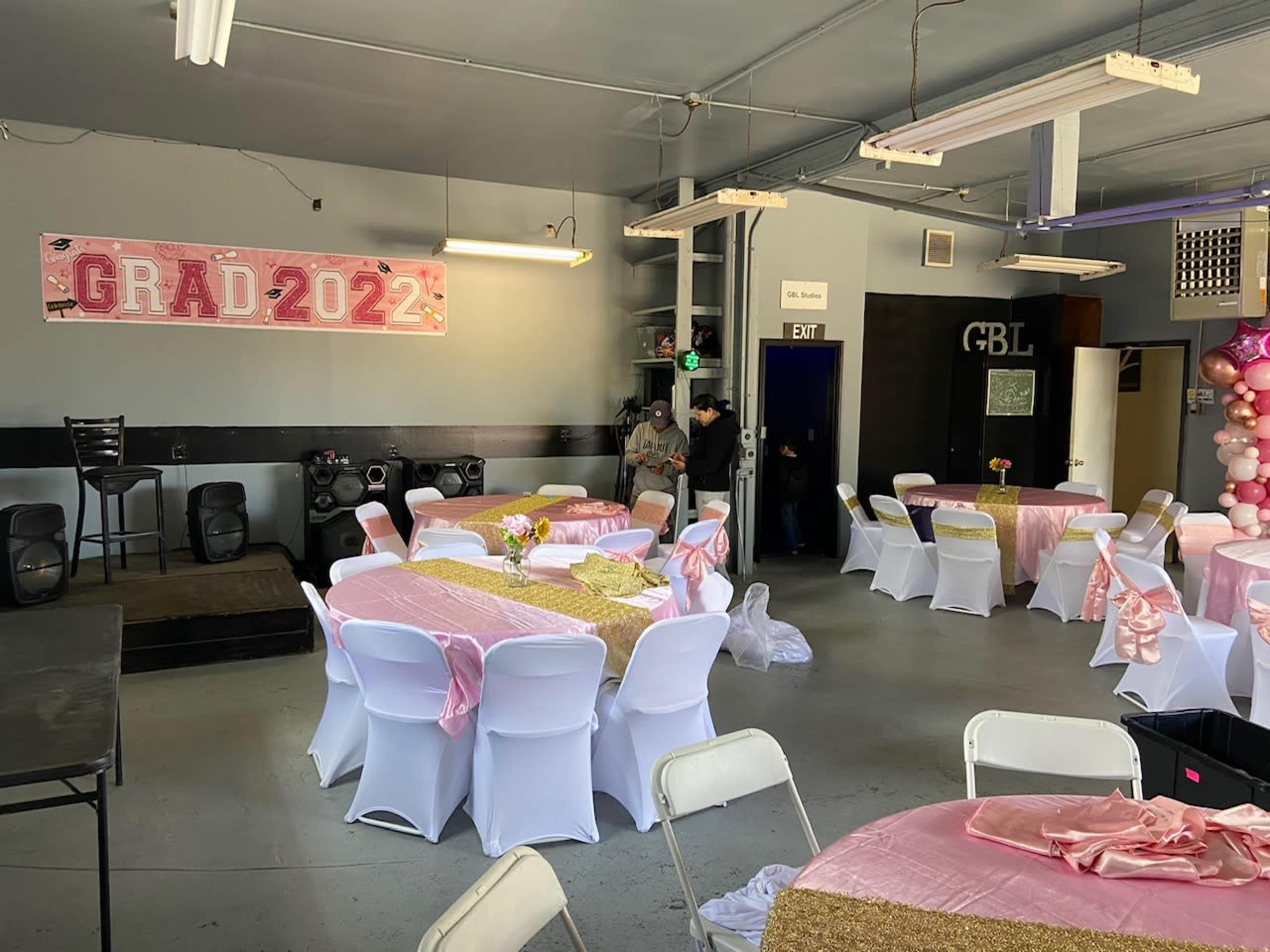 The image shows a decorated event space set up for a graduation celebration, featuring tables with pink decorations and a banner that reads "GRAD 2022."