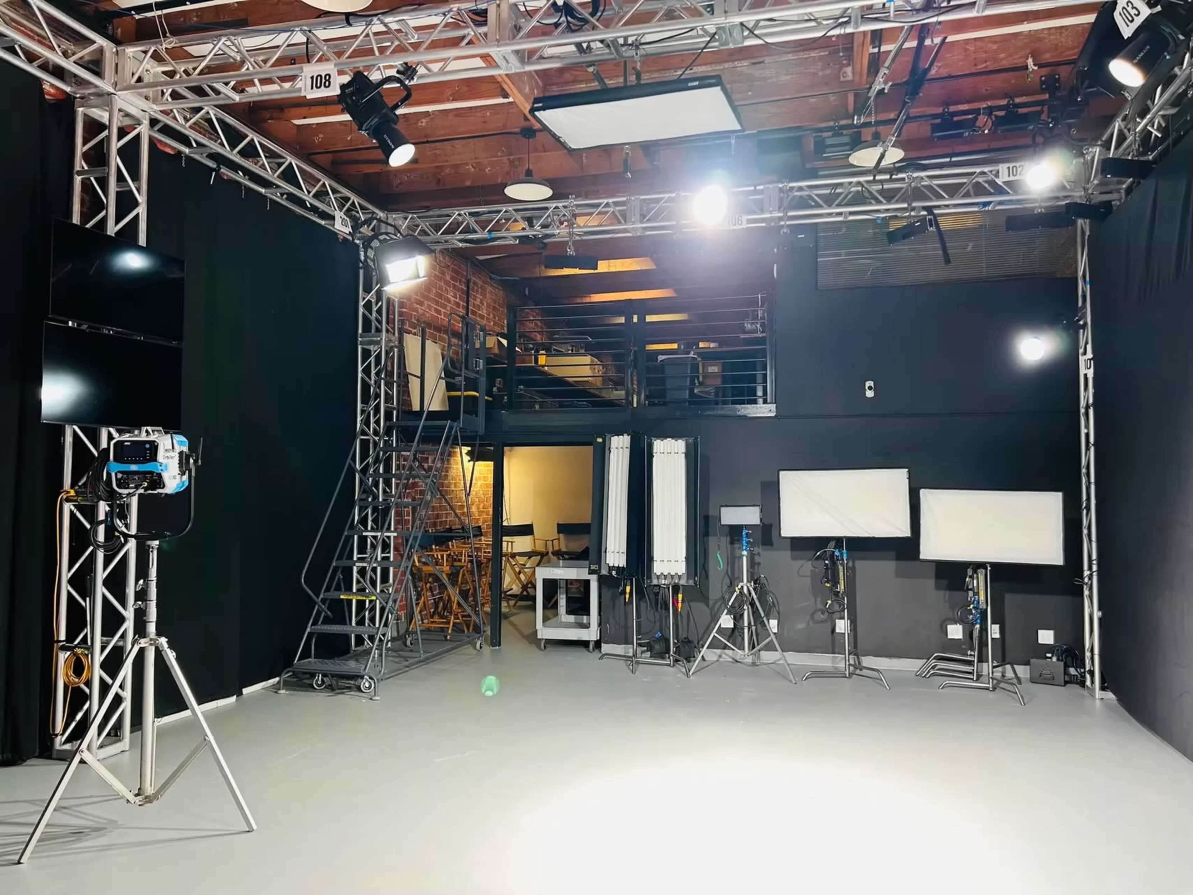 The image shows a well-lit studio space equipped with various lighting and camera equipment, black curtains, and a staircase in the background.