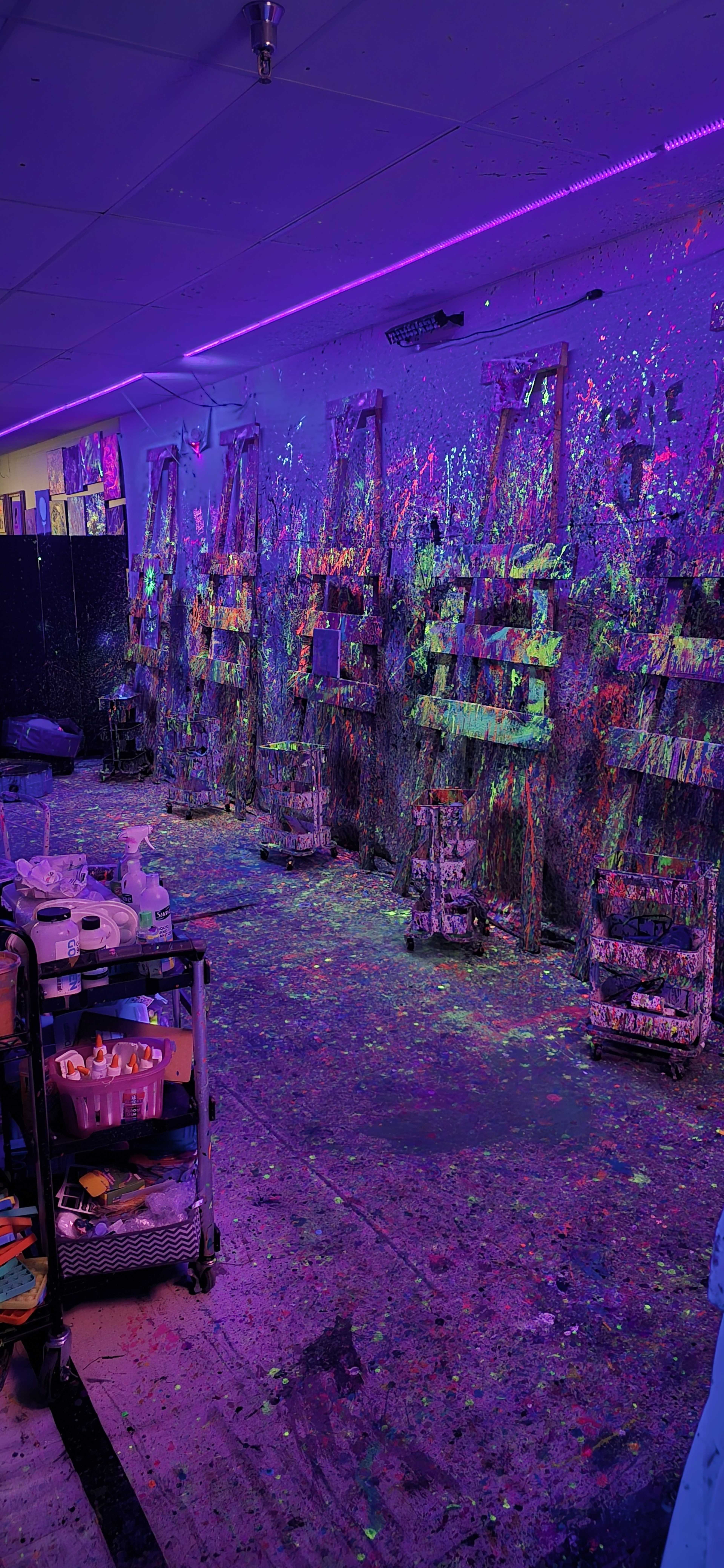 The image shows a vibrant art studio with colorful splatters on the walls and floor, featuring several easels lined up against a backdrop of UV lighting.