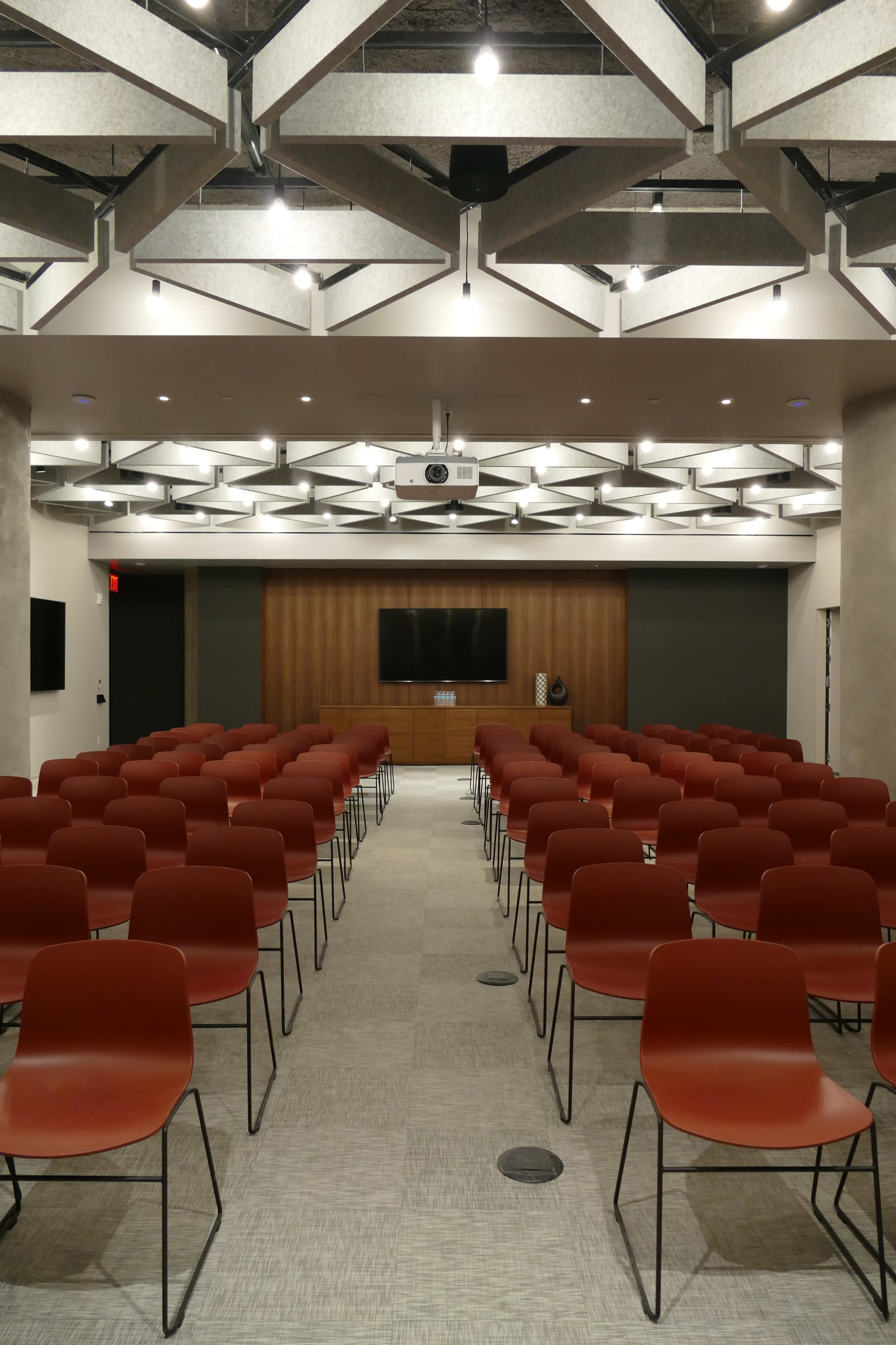 A modern conference room with rows of red chairs facing a wooden panel and a screen.