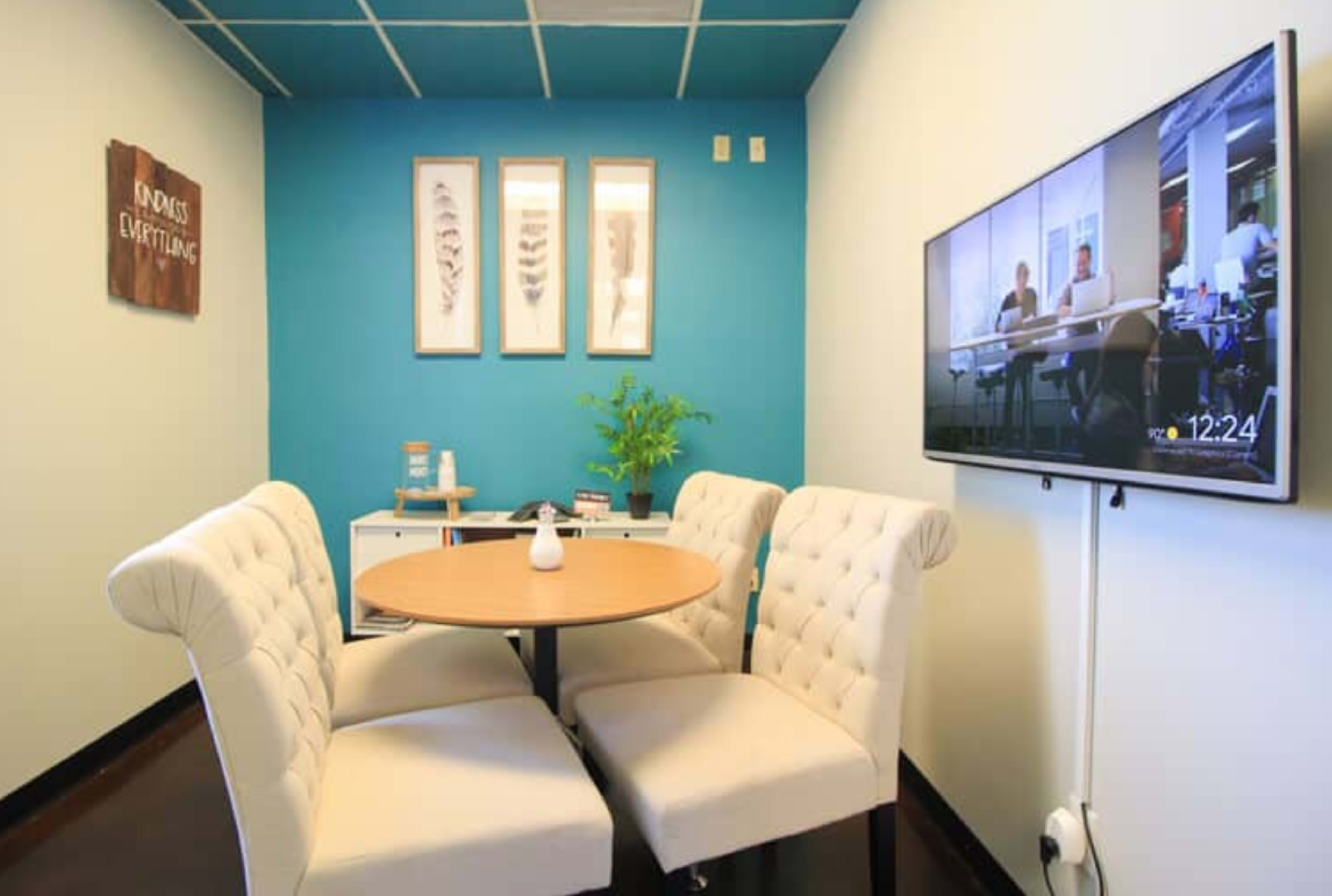 A small meeting area features a round table with four tufted chairs and a television mounted on the wall, alongside a plant and framed wall art.
