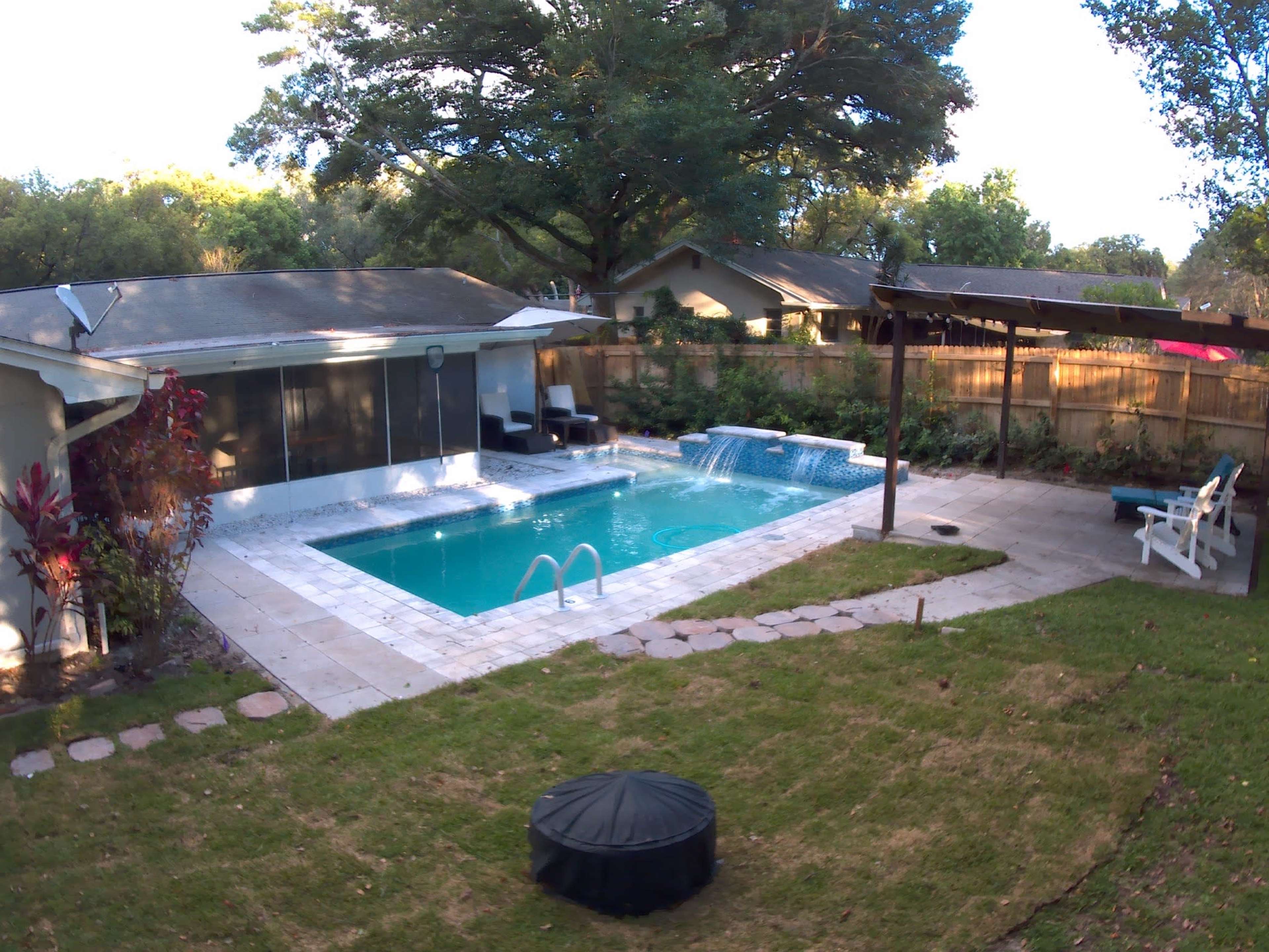 The image shows a backyard with a swimming pool, a covered patio area, and a lawn surrounded by trees and decorative landscaping.