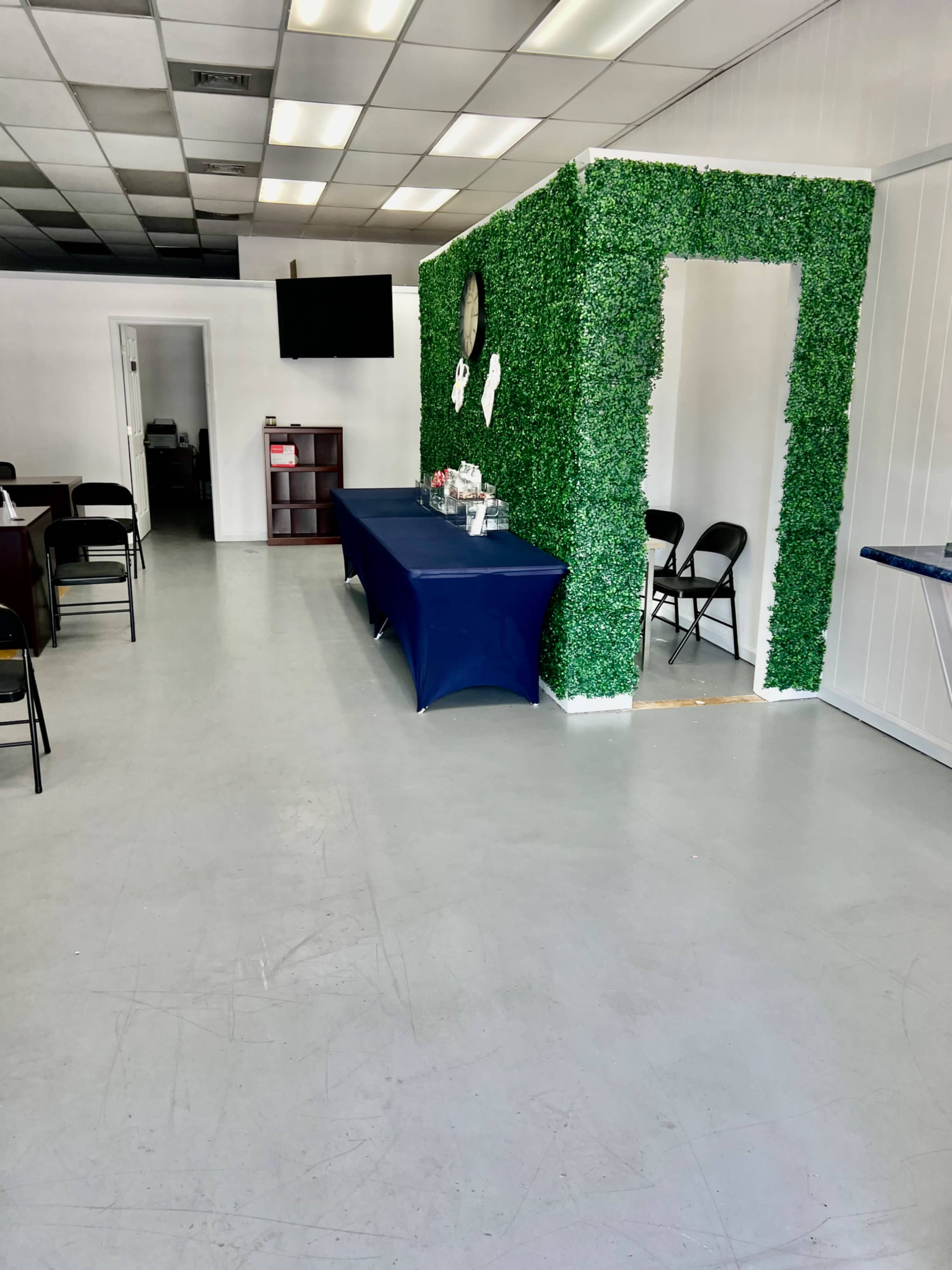 A room with a green, leafy wall structure, a table covered with a blue cloth, and black chairs arranged around it, with a doorway leading to another space.