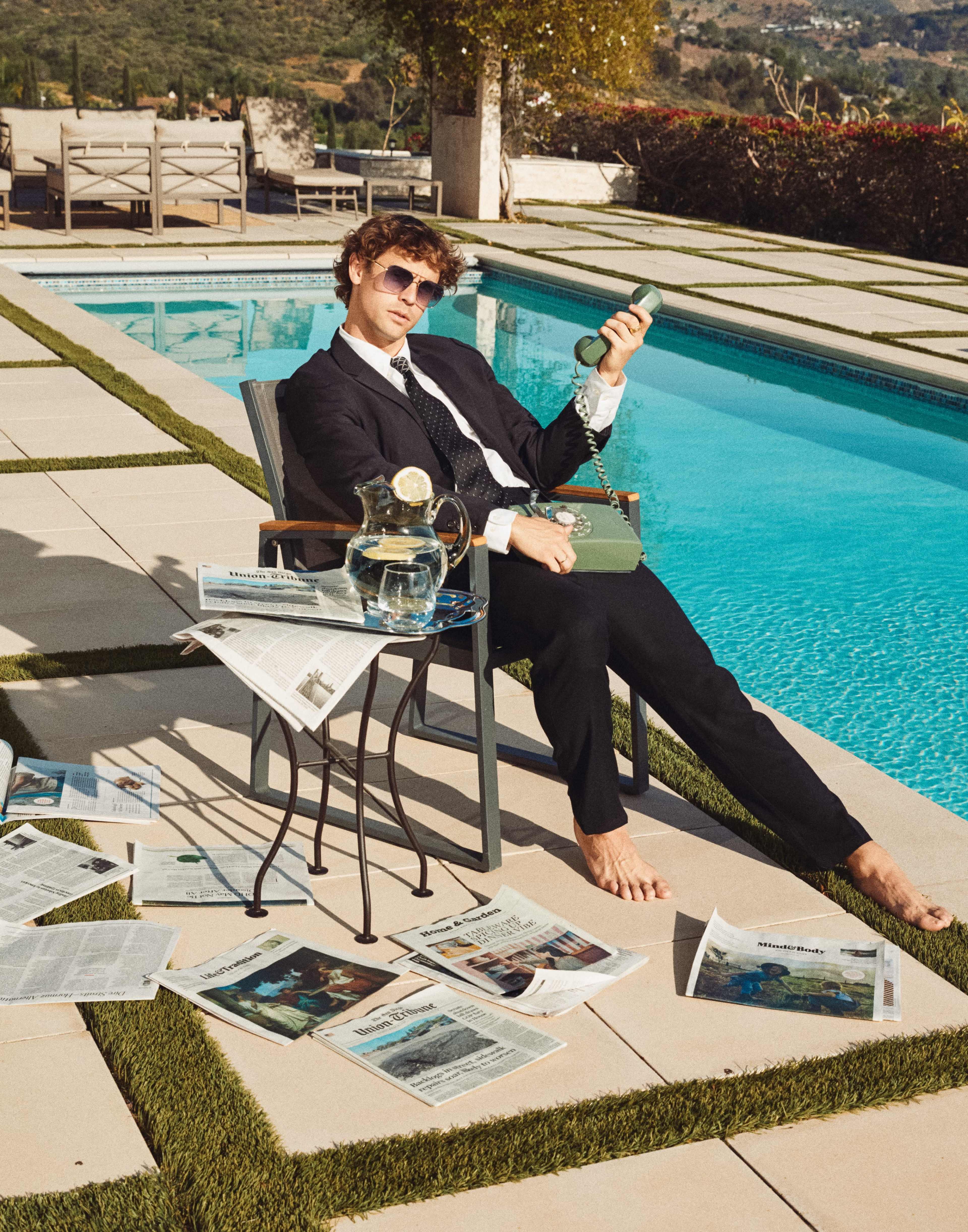 A man in a suit sits in a chair by a swimming pool, casually holding a vintage telephone, surrounded by scattered newspapers and a pitcher of water on a small table.