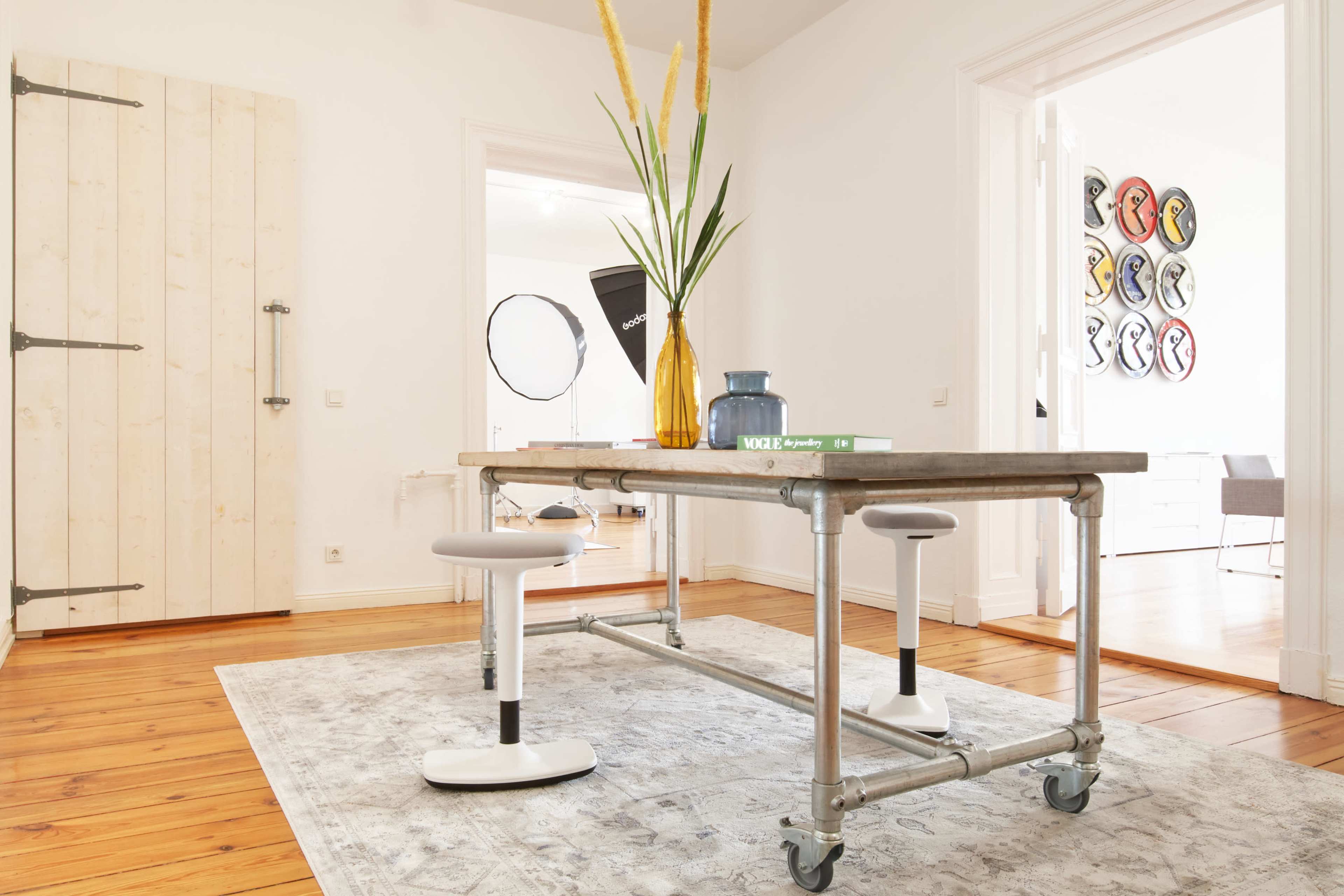 A minimalist room features a wooden table with metal legs, flanked by two stools, and adorned with a vase of tall plants and a stack of books.