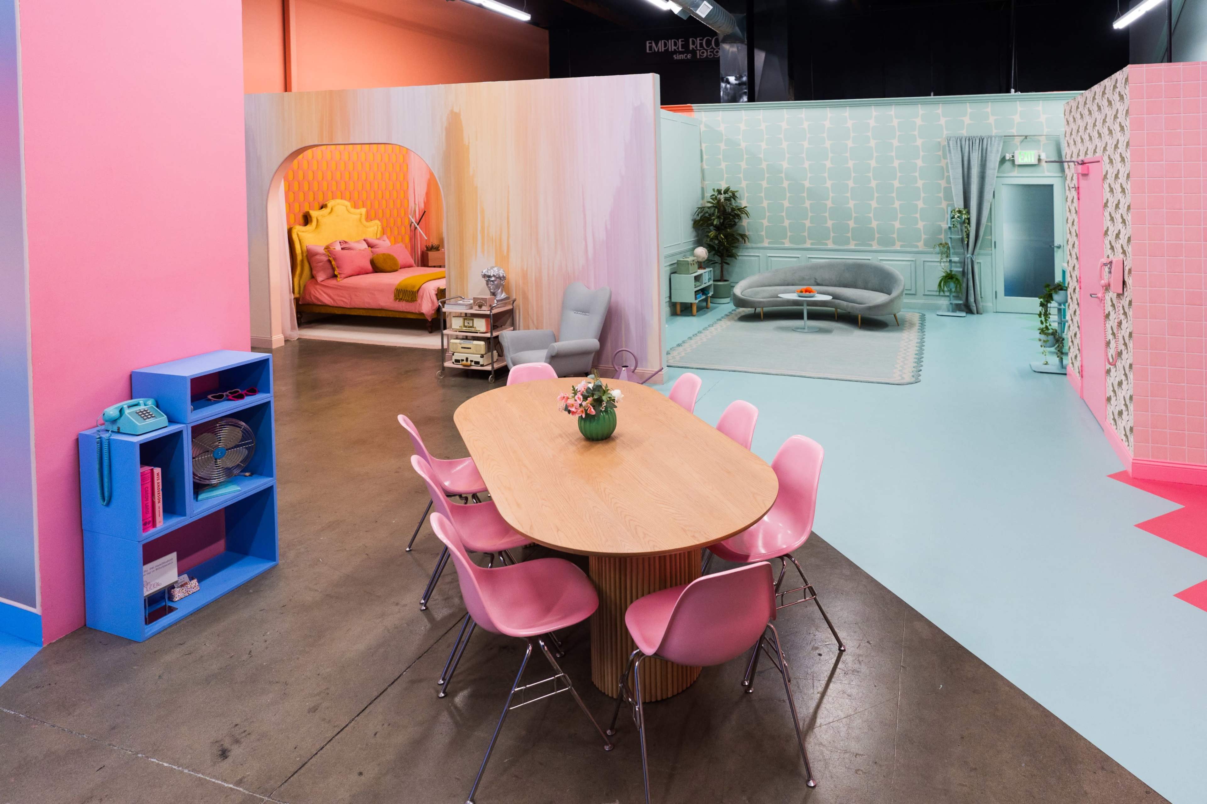 The image shows a brightly colored interior space divided into different styled sections, including a bedroom, living area, and dining area with a round table and pink chairs.