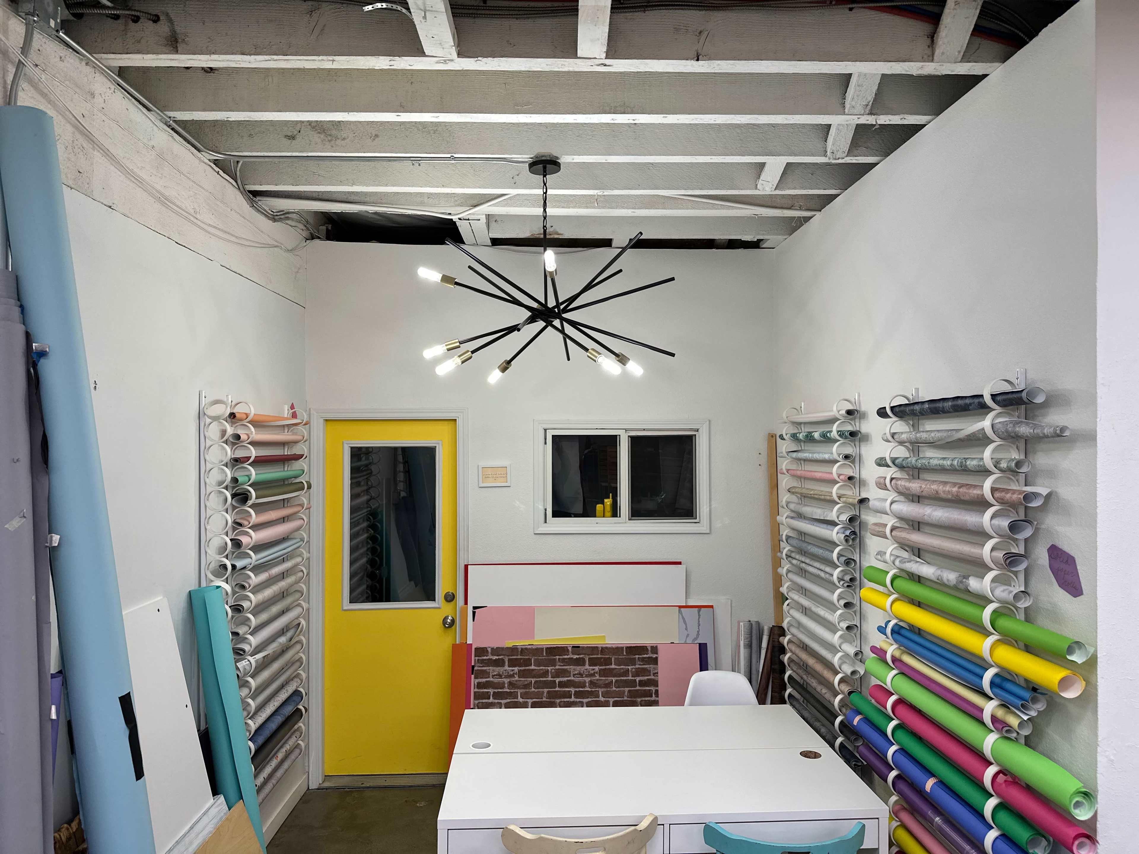 A well-lit craft room features colorful rolls of material organized on wall-mounted racks, with a bright yellow door and a minimalist chandelier.