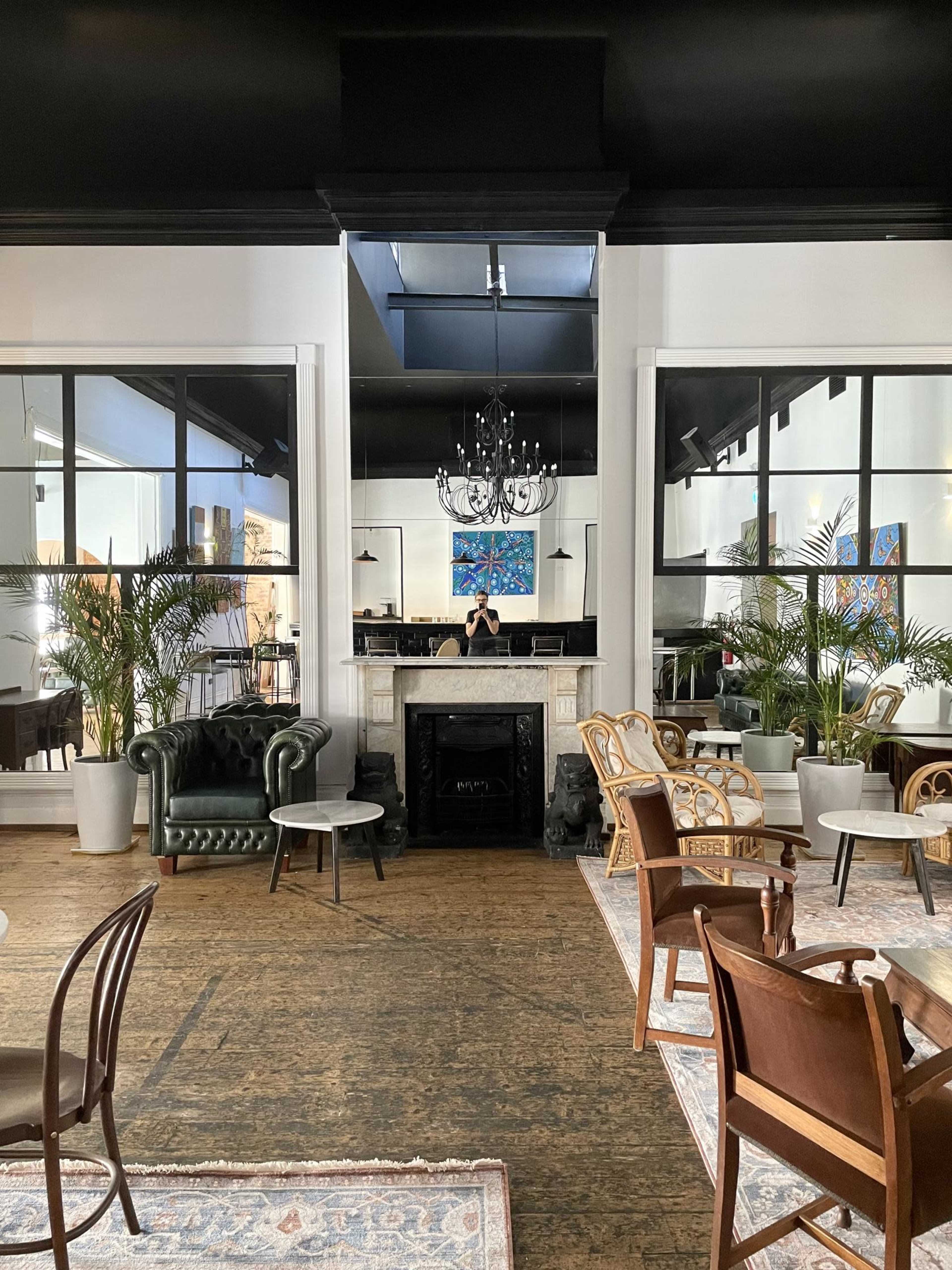 The image shows a stylish indoor café or lounge with an arrangement of chairs and small tables, large windows, plants, and a fireplace.