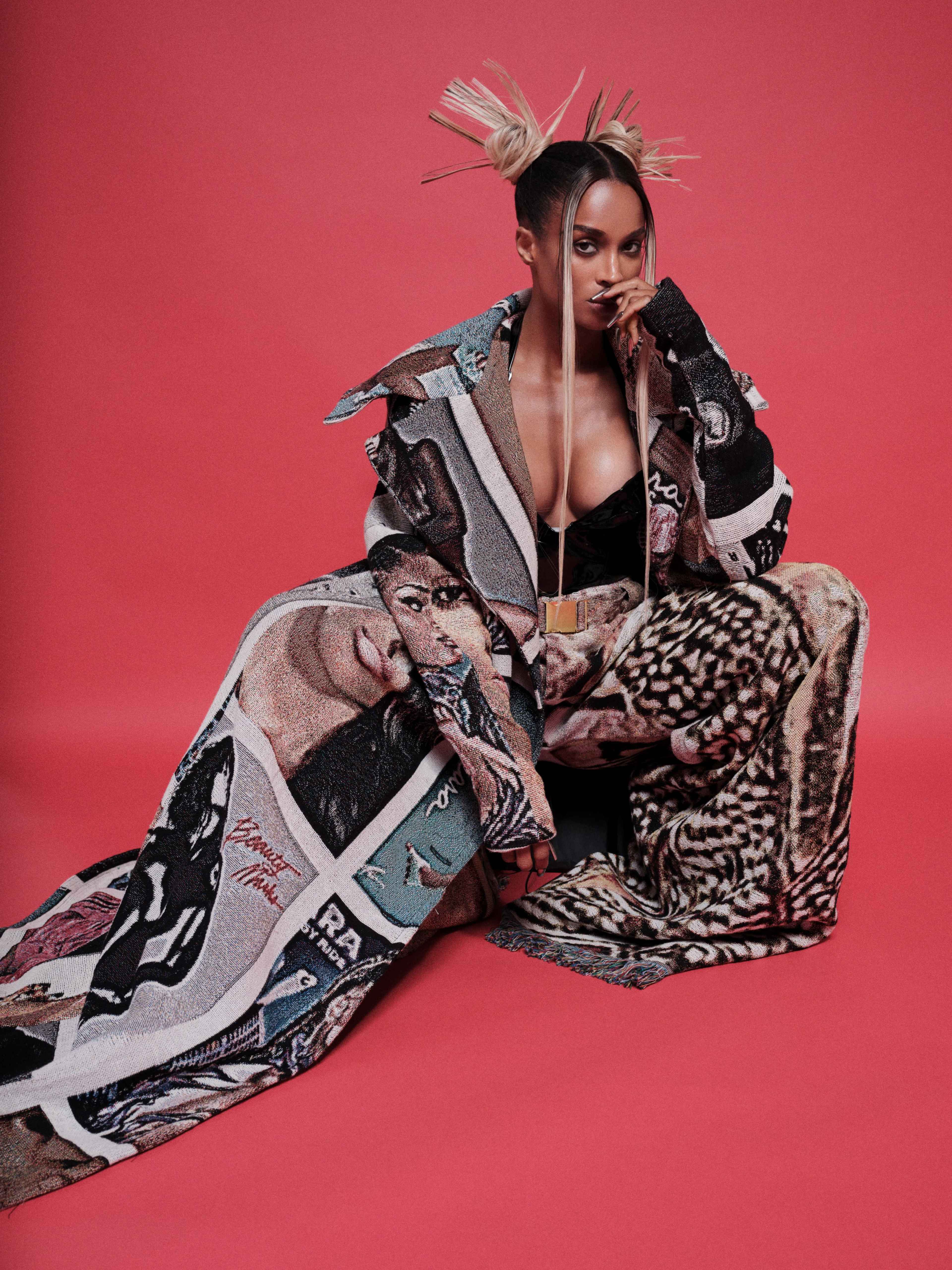 A person sits on the floor, wrapped in a patterned blanket, against a bright red background, with unique hair accessories and bold clothing.
