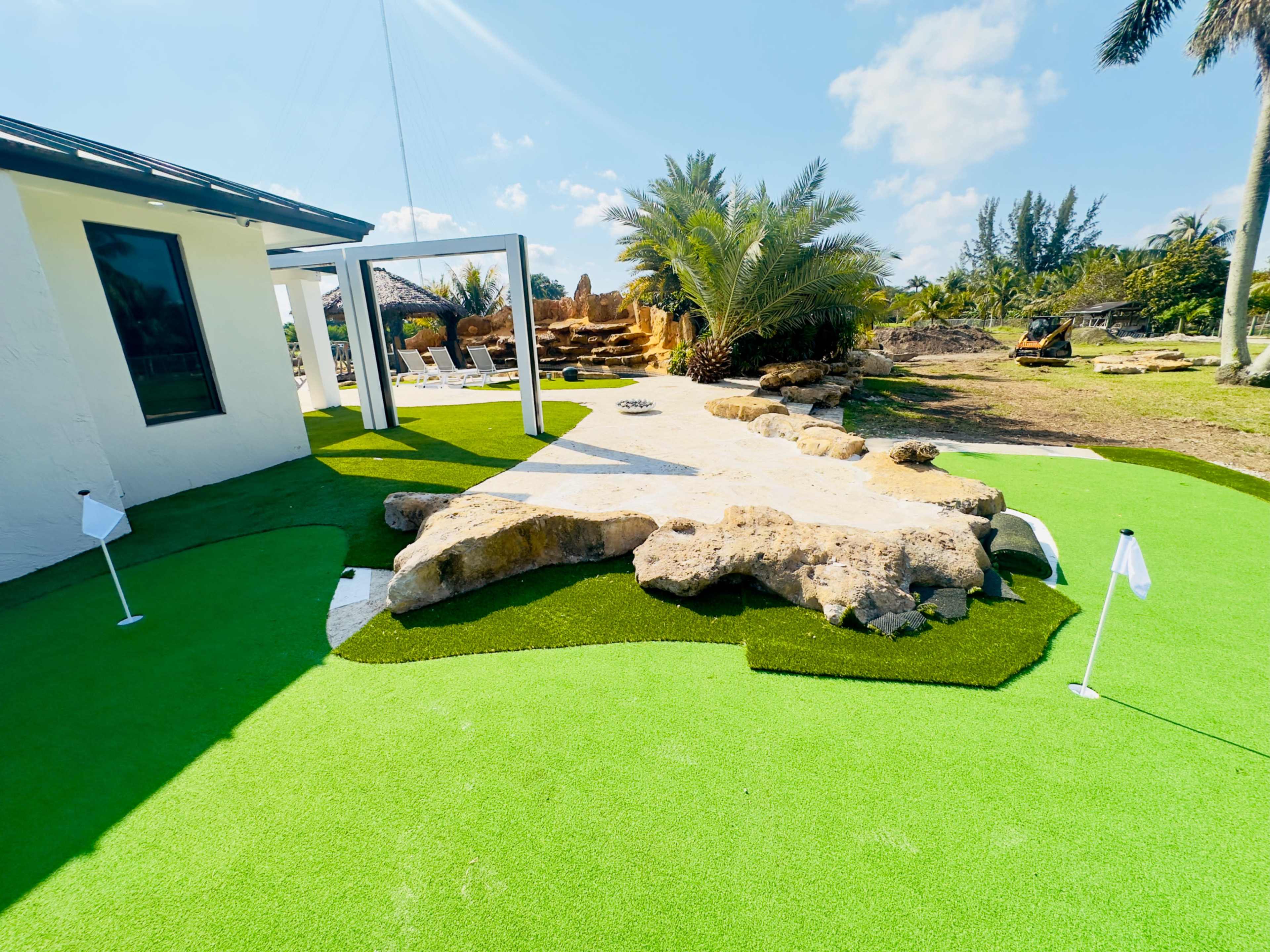 The image shows a landscaped outdoor area featuring artificial grass, stones, and a pathway, surrounded by palm trees and open space.
