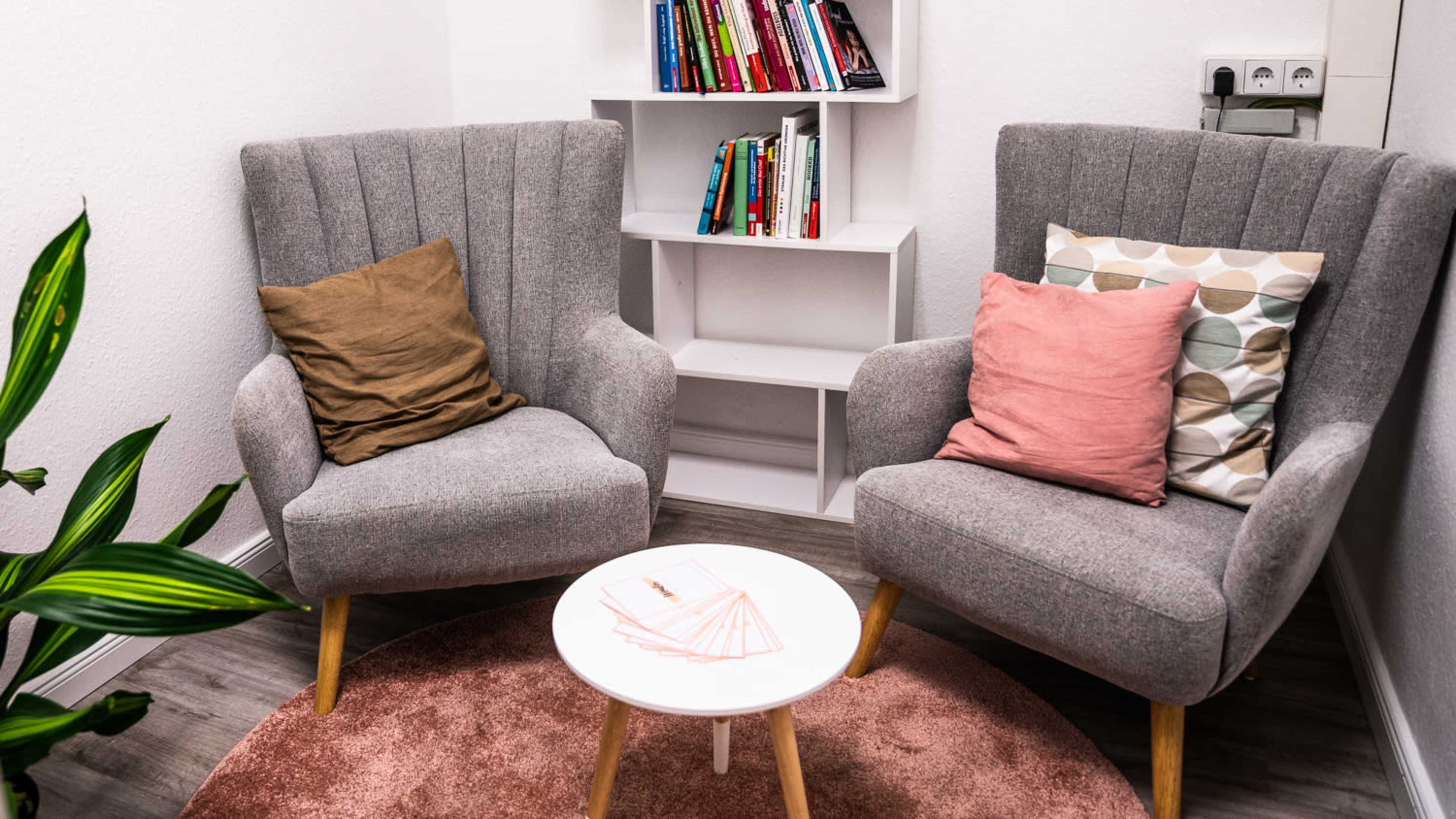Two gray armchairs with assorted pillows positioned around a small round table on a rug, with a bookshelf filled with colorful books in the background.