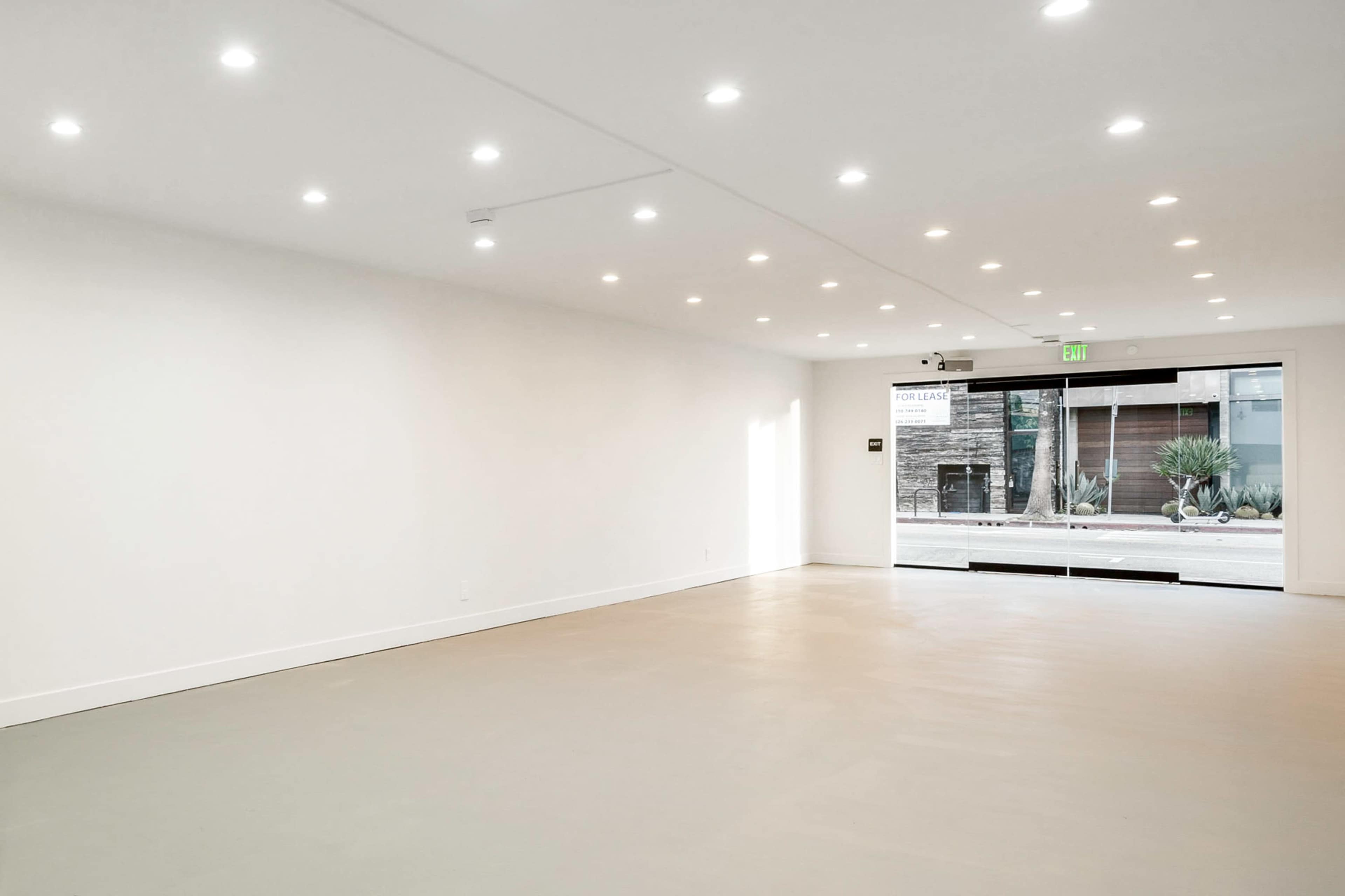 An empty room with a smooth grey floor, white walls, and bright overhead lights, featuring large glass doors that open to a view of the exterior.