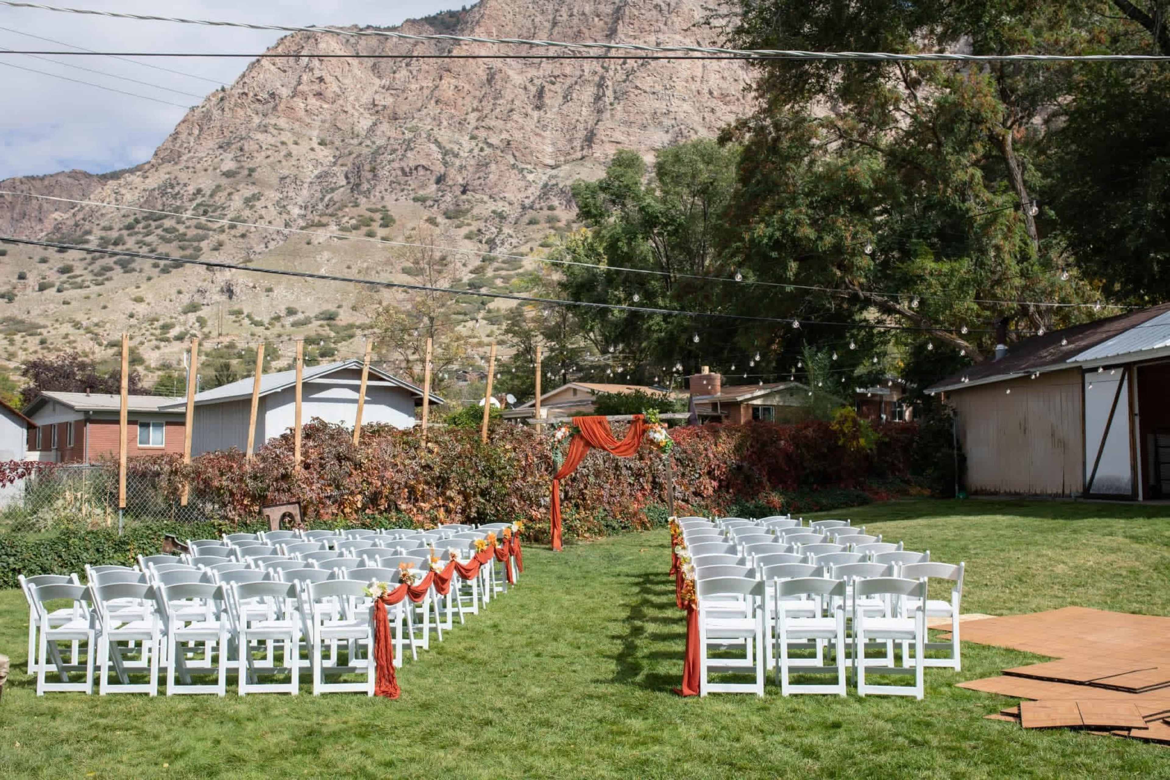 A grassy outdoor area is set up for an event with rows of white chairs facing a decorated arch under a backdrop of mountains.