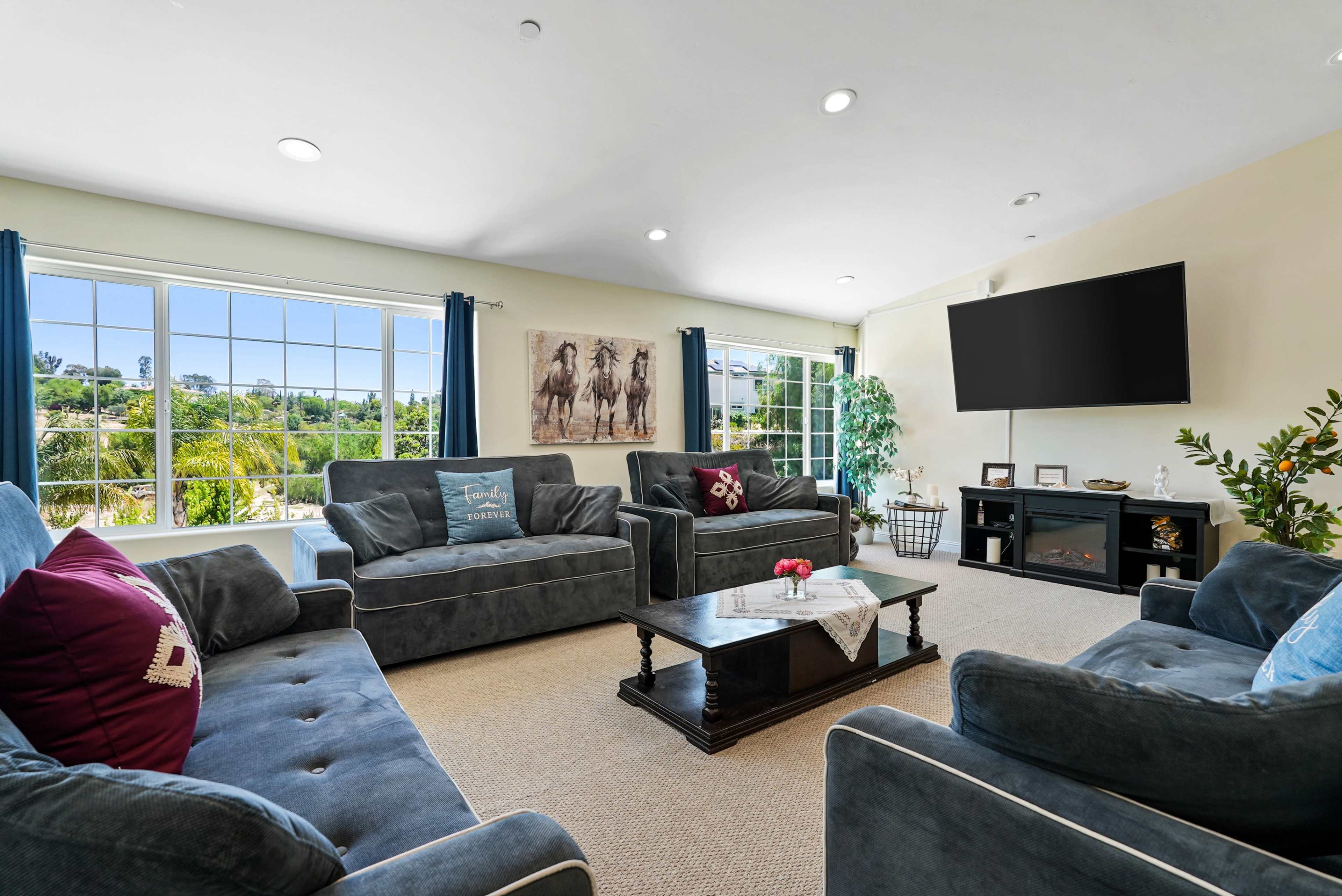 The living room features gray sofas arranged around a central table, with large windows providing a view of greenery outside and a wall-mounted television.