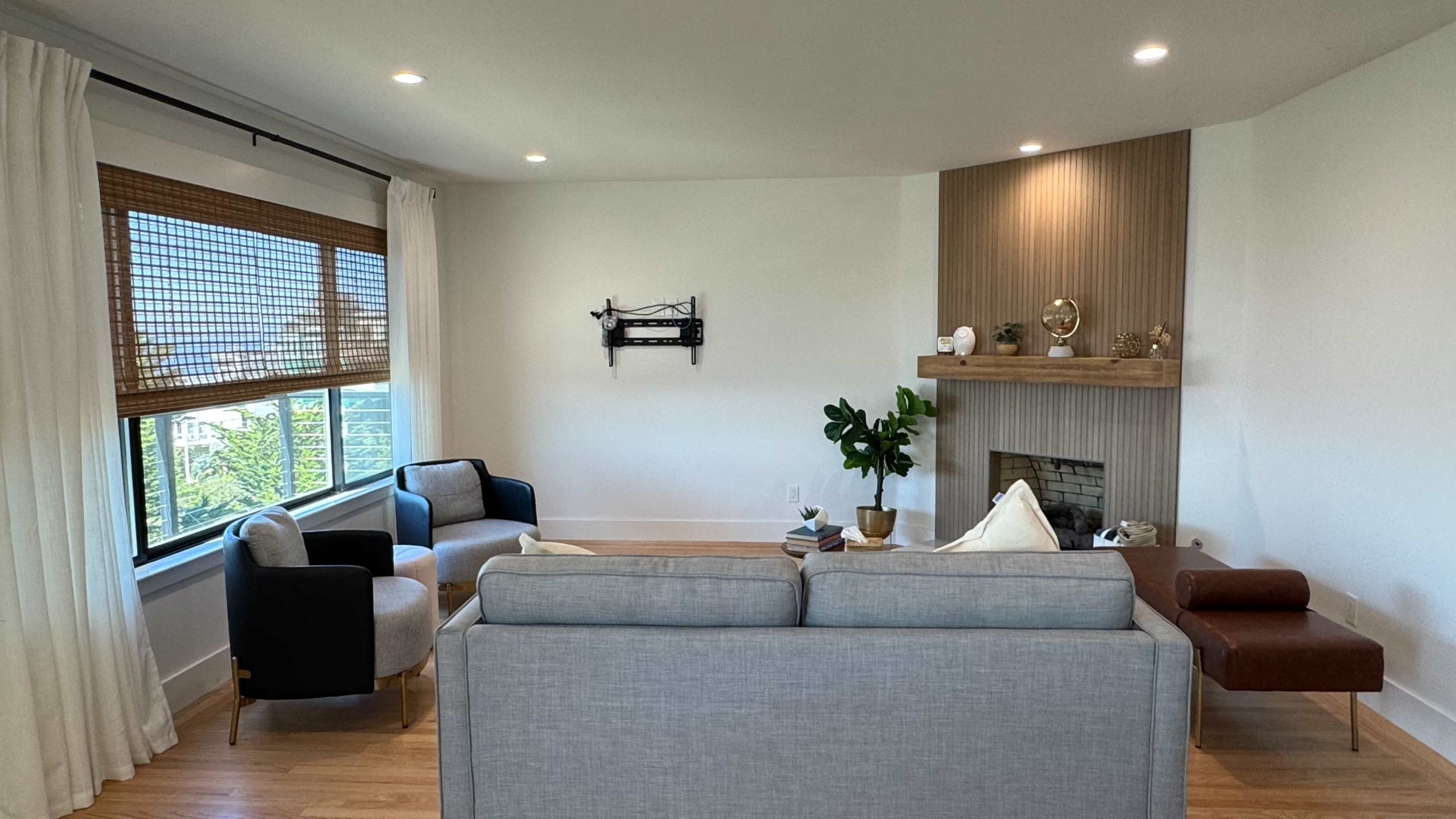 The image shows a modern living room featuring a gray sofa, two armchairs, a wooden mantel with decor, and a large window with natural light.