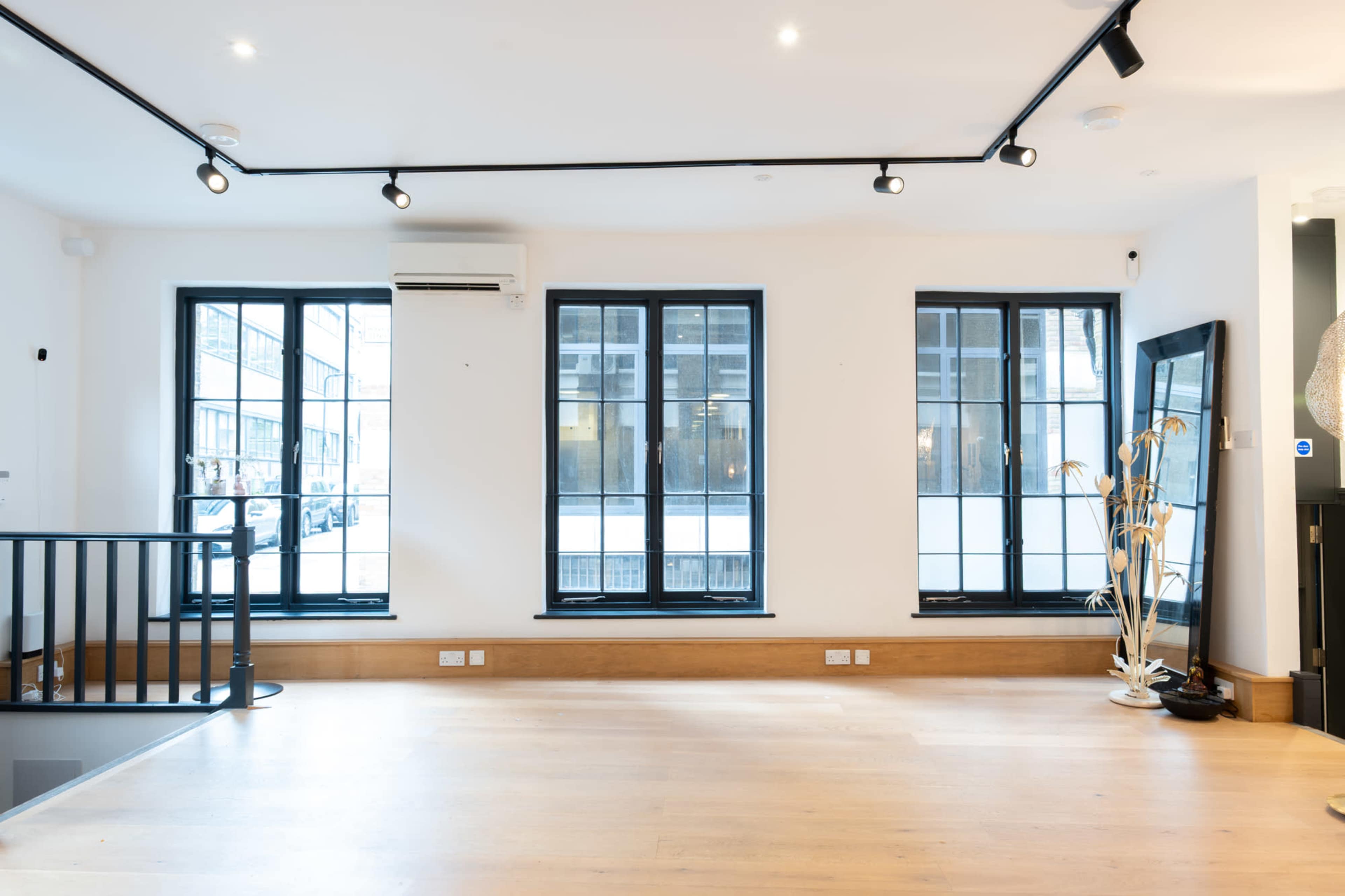 The image shows a bright, open room with three large black-framed windows and wooden flooring.