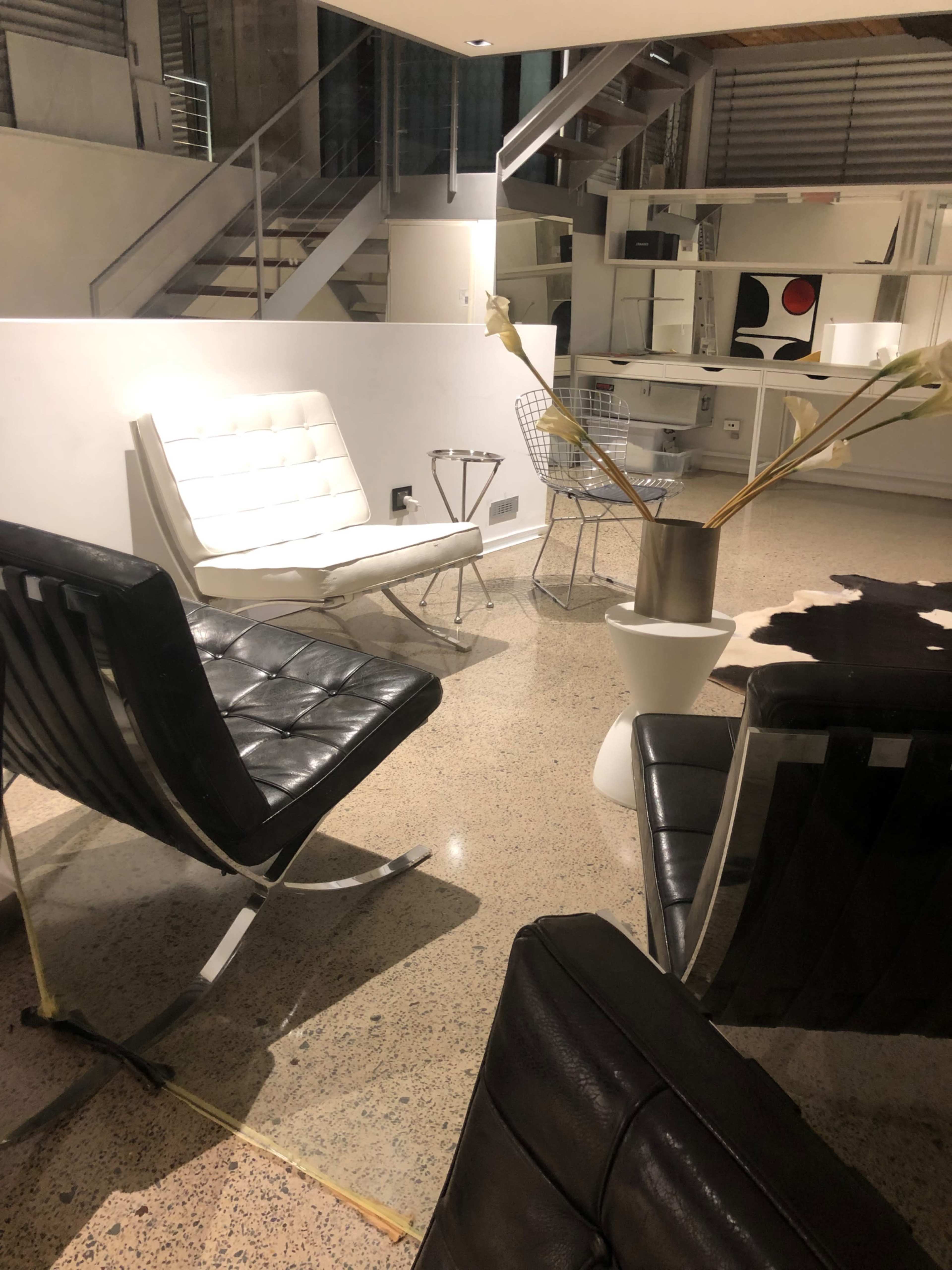 The image shows a modern indoor living space with minimalist furniture, including black and white chairs, a small table with a vase, and a staircase in the background.
