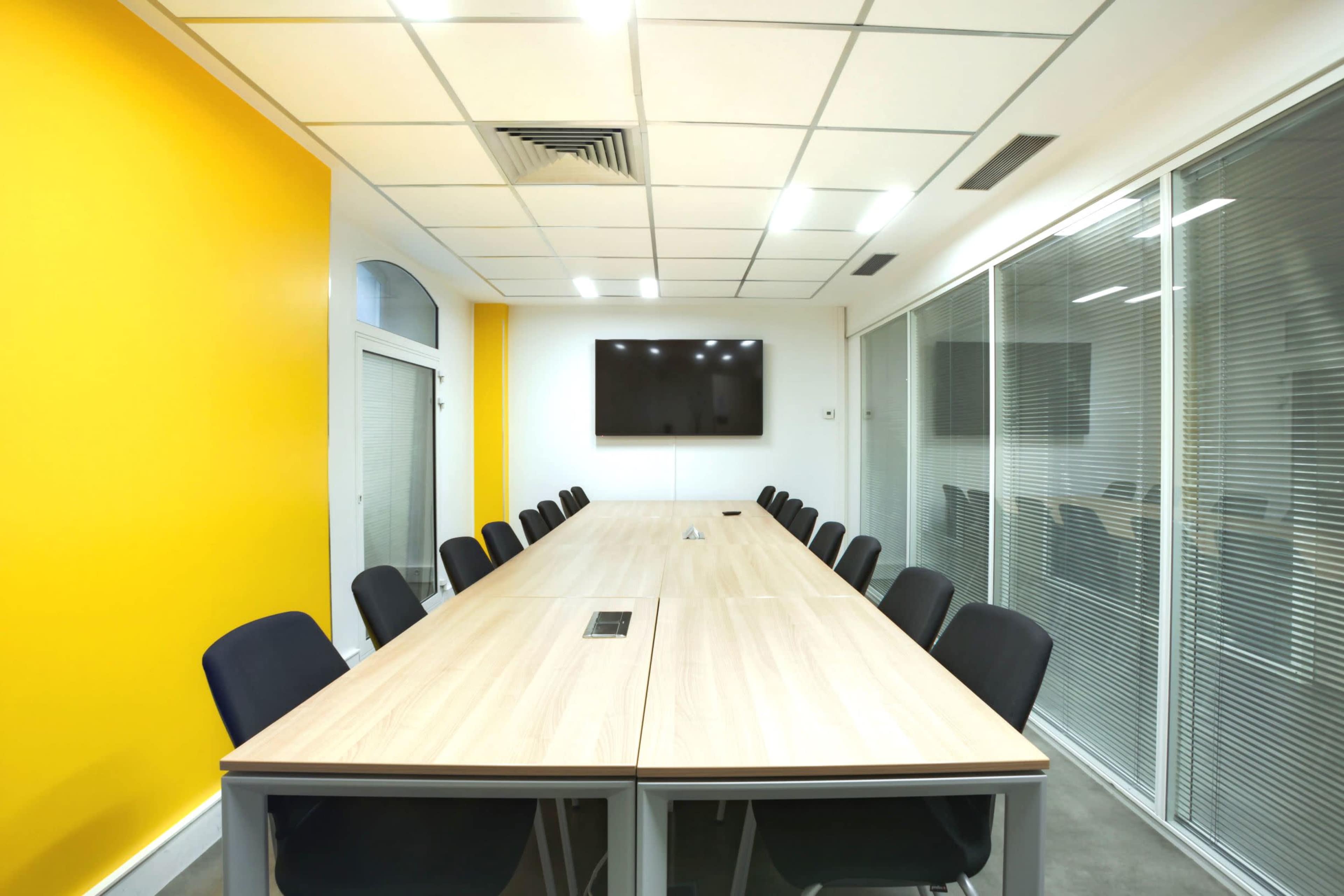 A modern conference room features a long wooden table surrounded by black chairs, with a large television mounted on the wall and a bright yellow accent wall.