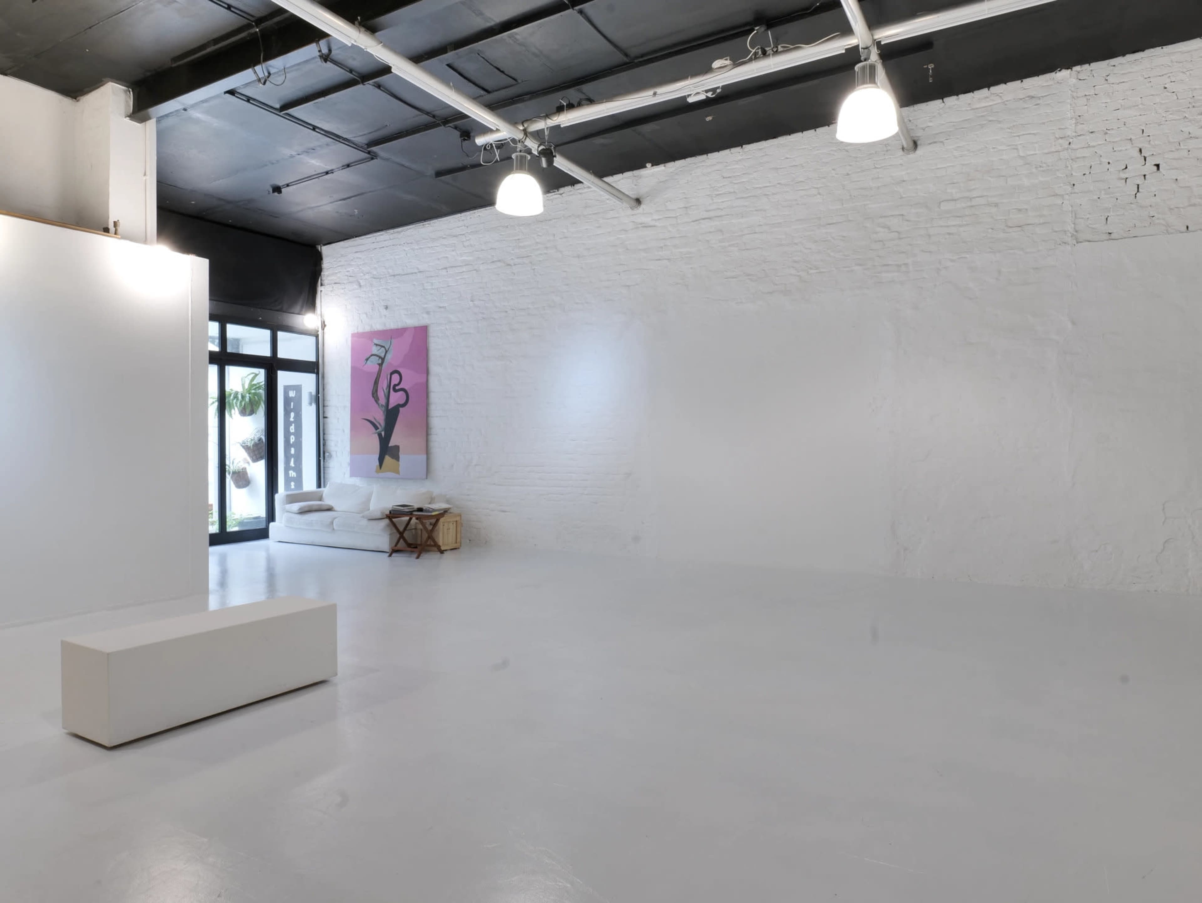 The image shows a spacious, minimalist interior of a gallery with white walls, a bench in the center, and a single piece of artwork on the wall.