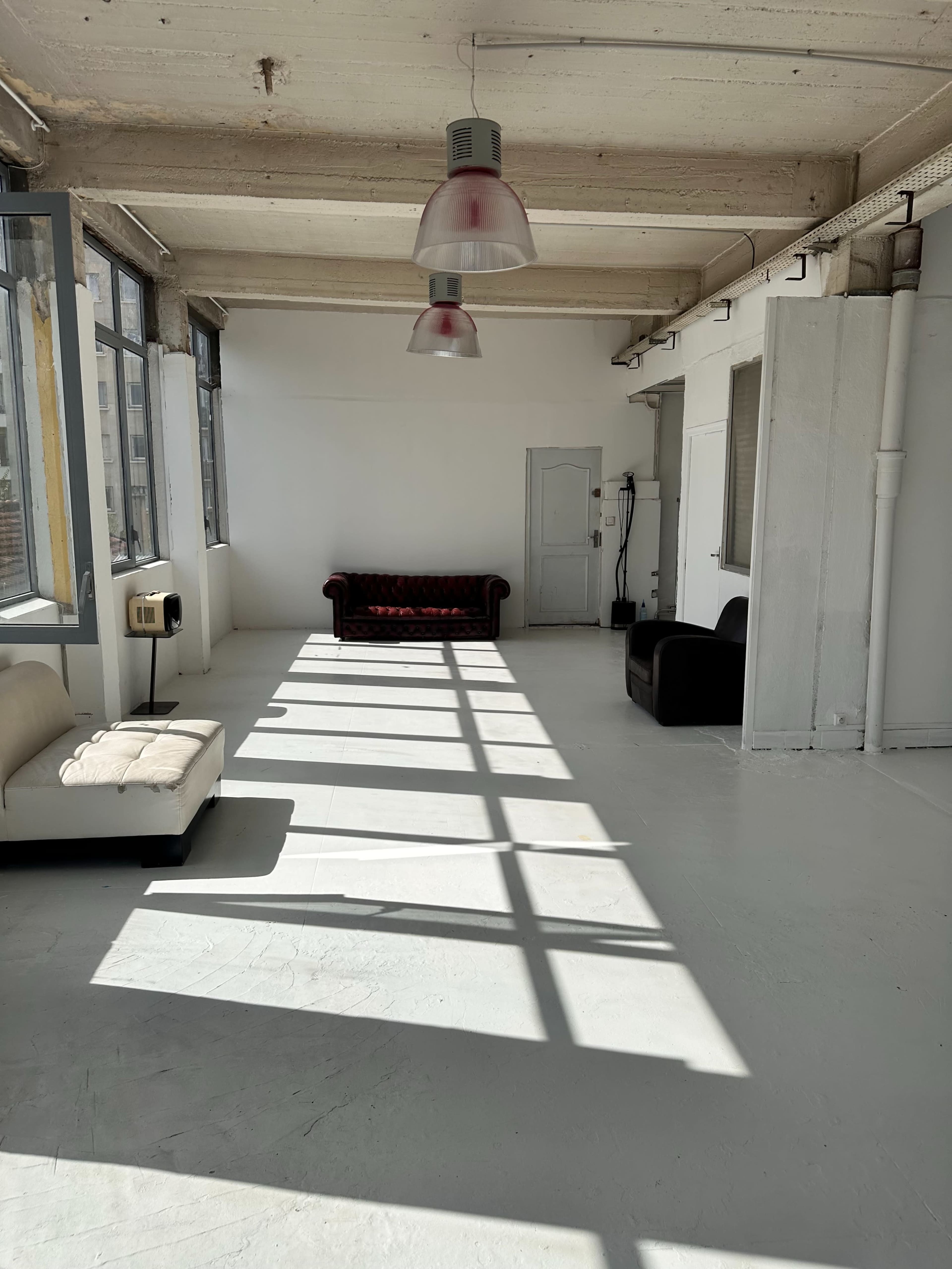 The image shows a spacious, minimalistic room with a white floor, large windows, and two sofas casting long shadows in sunlight.