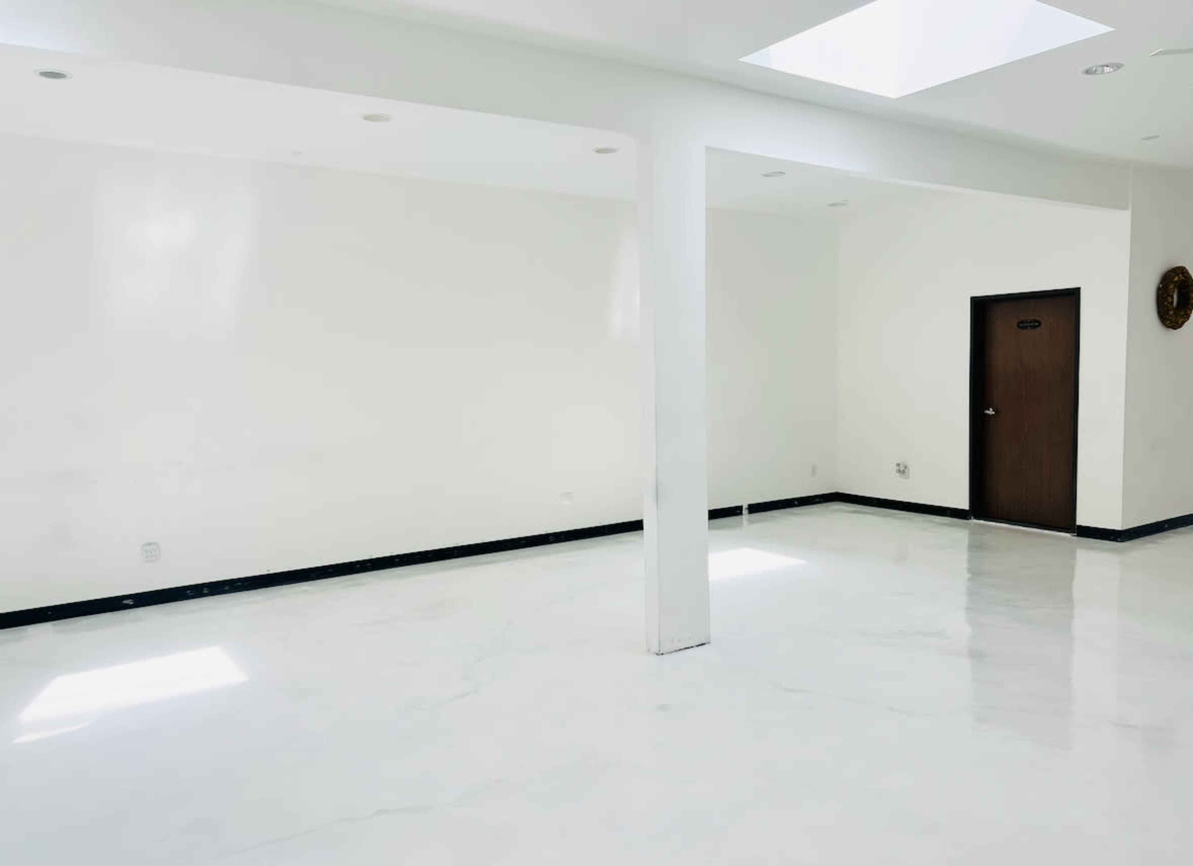 The image shows a spacious, empty room with white walls and a glossy, white floor, featuring a skylight and a single door in one corner.