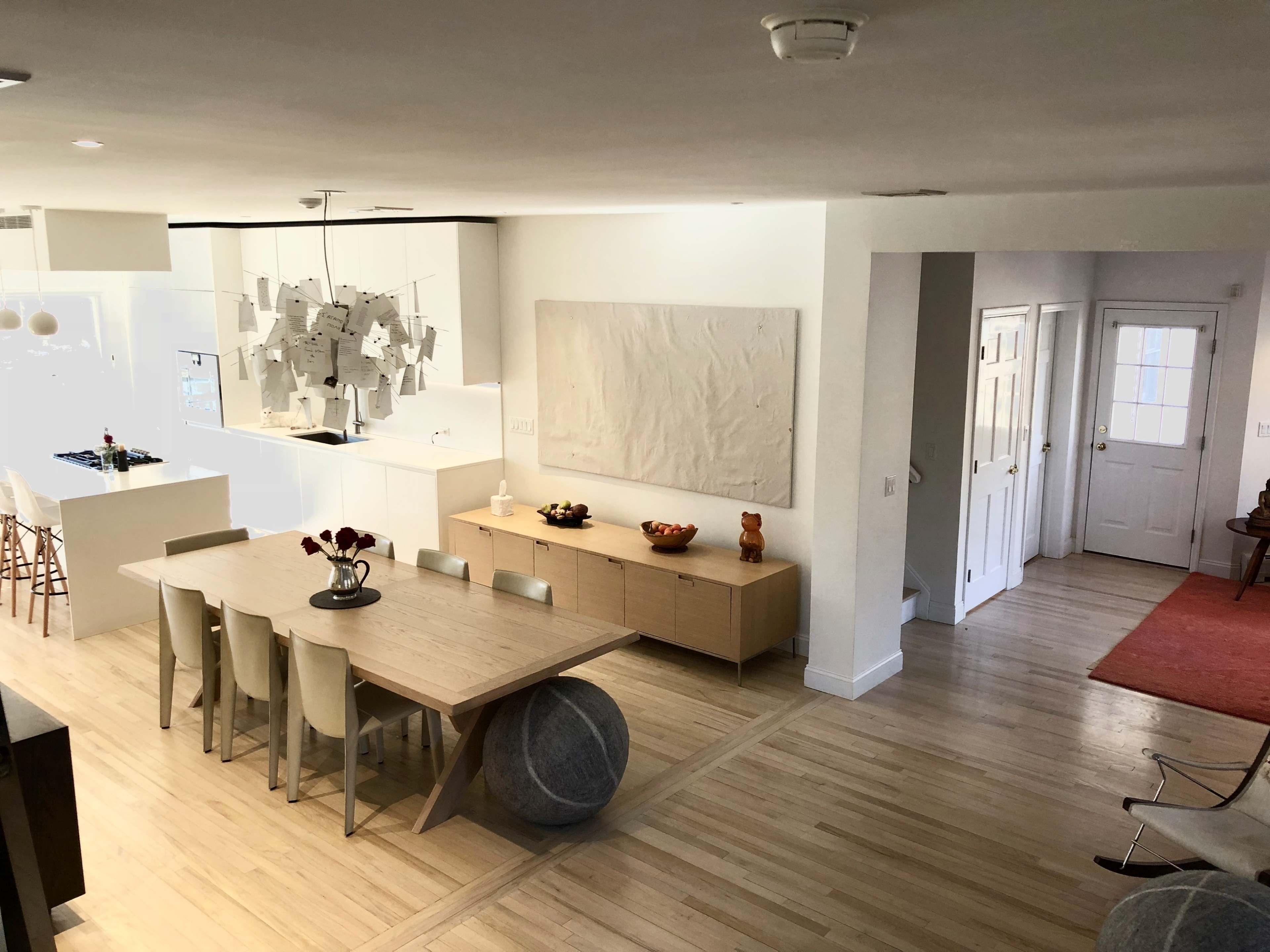 A modern, open-concept living space featuring a dining area with a large table, minimalist decor, and a view of a kitchen and entryway.