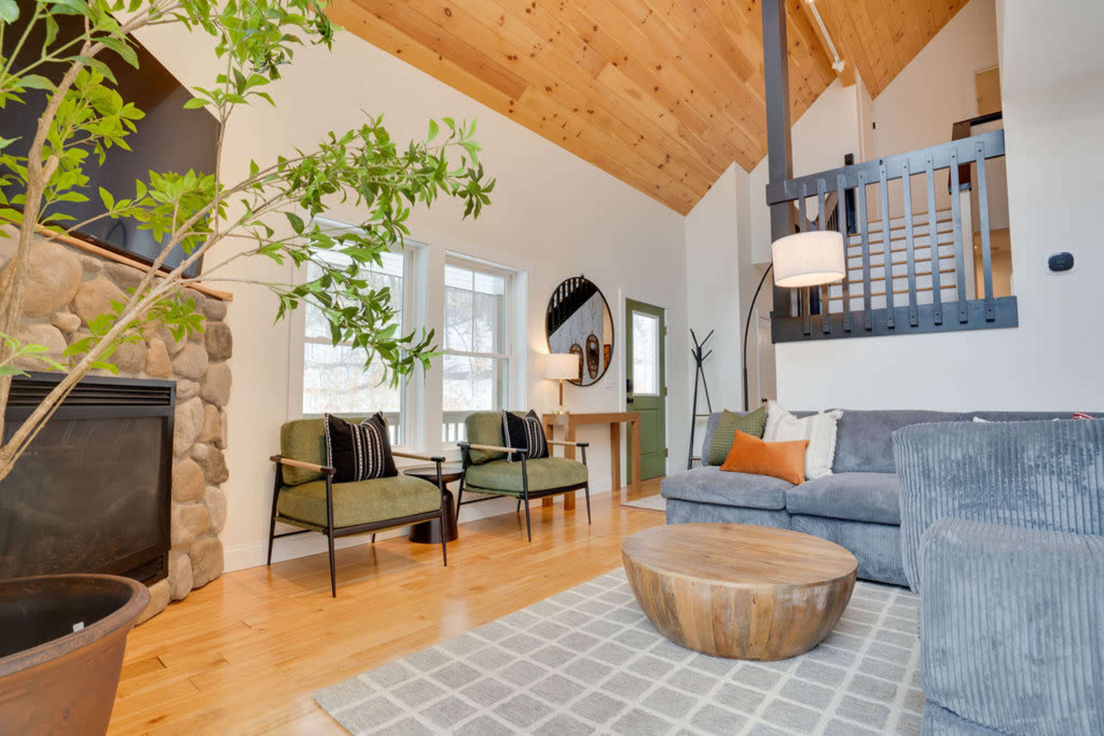 The living room features a stone fireplace, two green chairs, a gray sofa, a round coffee table, and a staircase in the background.