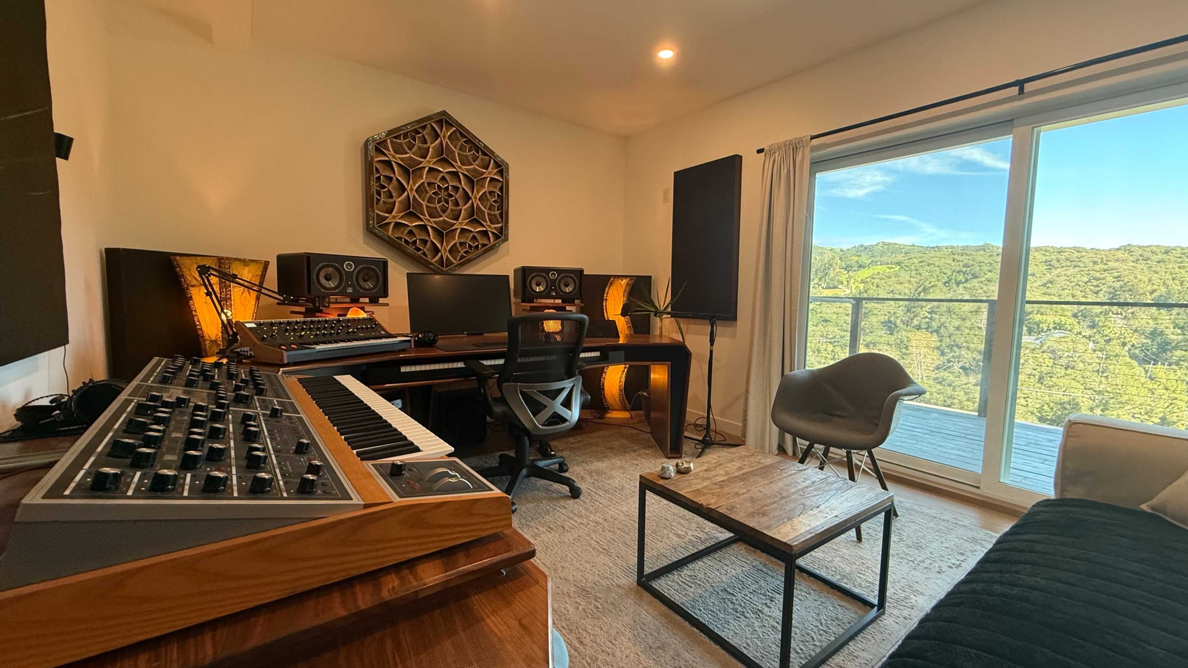 A modern home studio featuring a synthesizer, computer setup, and large windows overlooking a forested landscape.