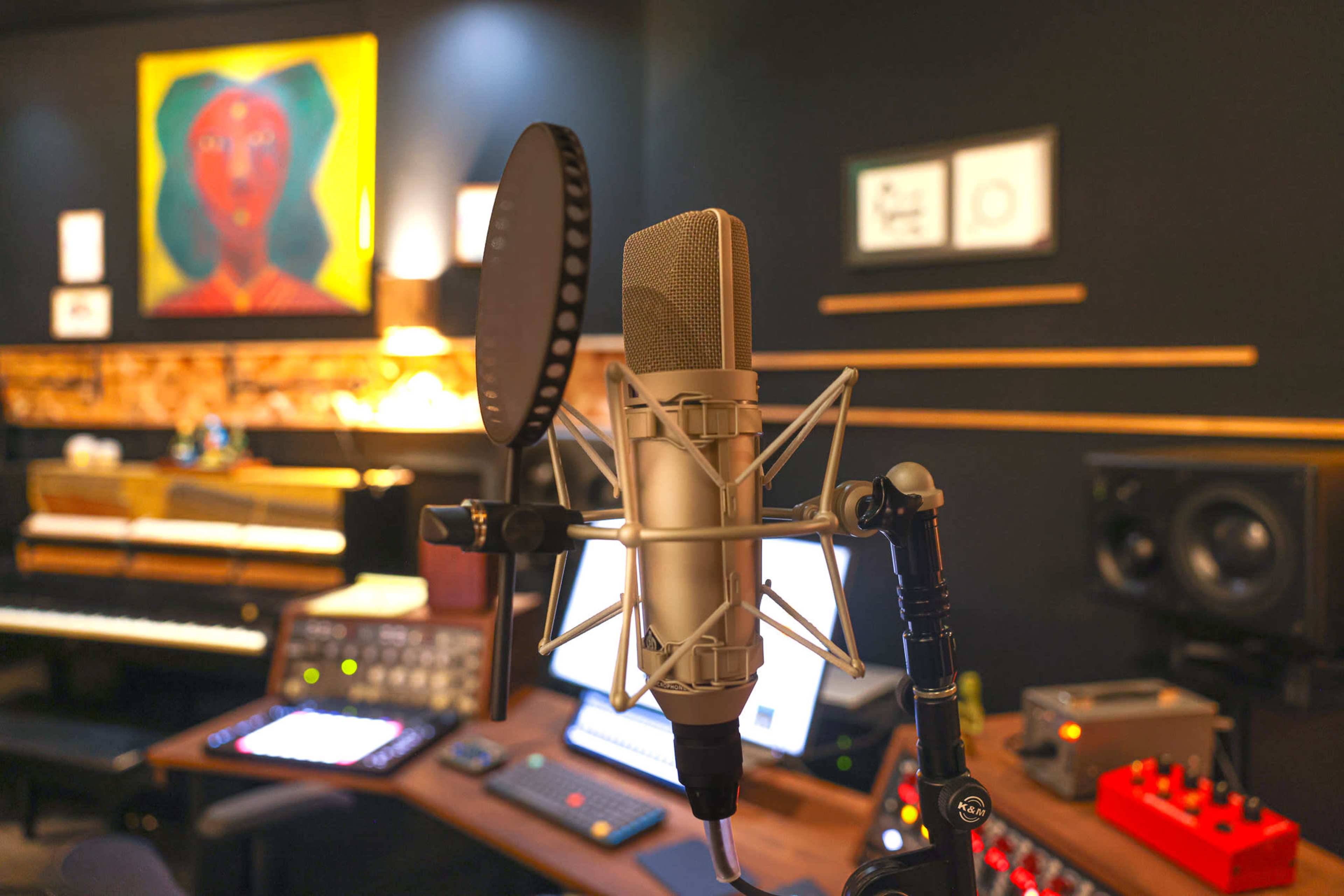 The image shows a close-up of a microphone in a music studio with various recording equipment and instruments in the background.