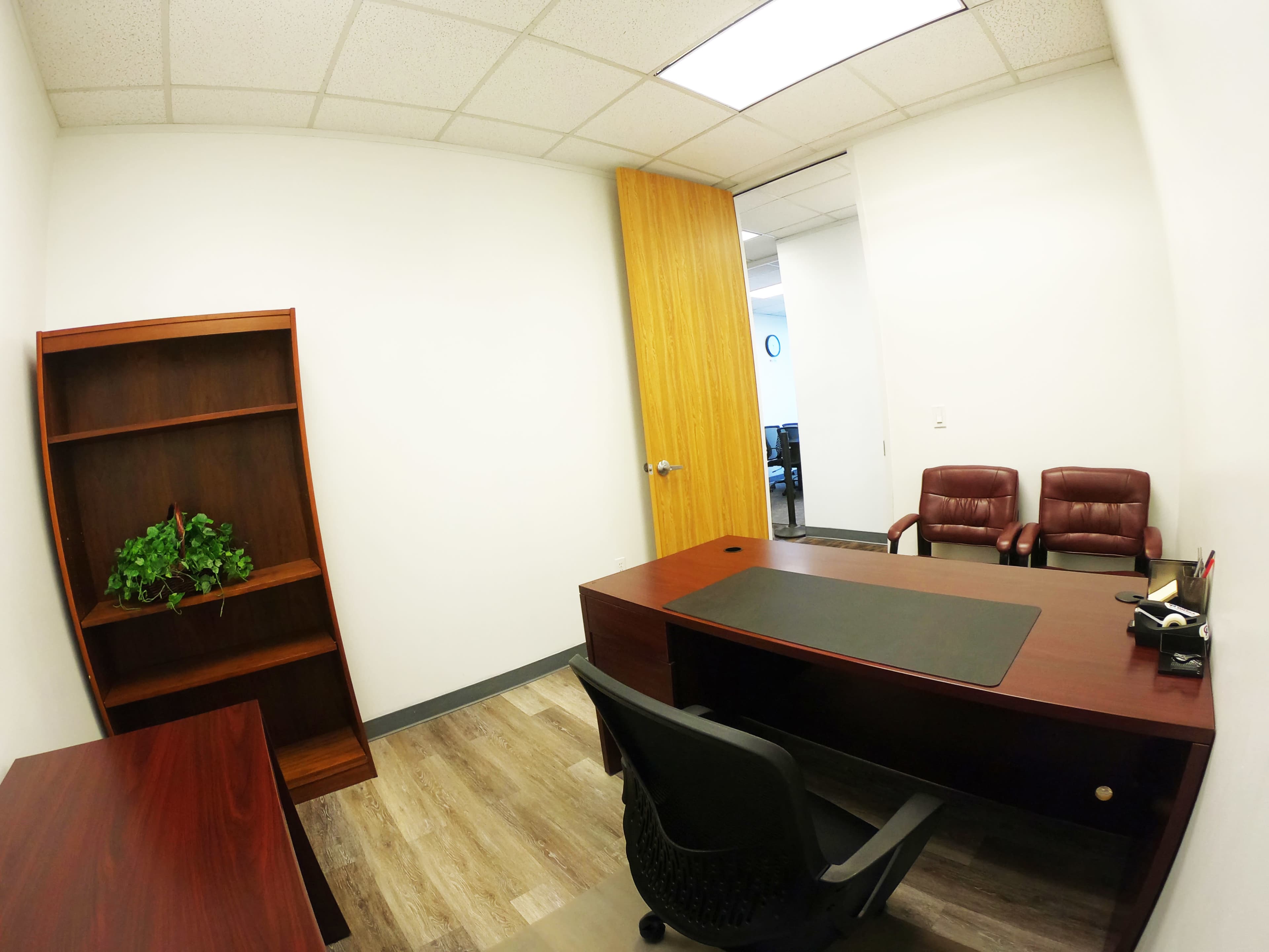 A small office space with a brown wooden desk, a chair, a bookshelf with a plant, and two brown leather chairs against a light-colored wall.