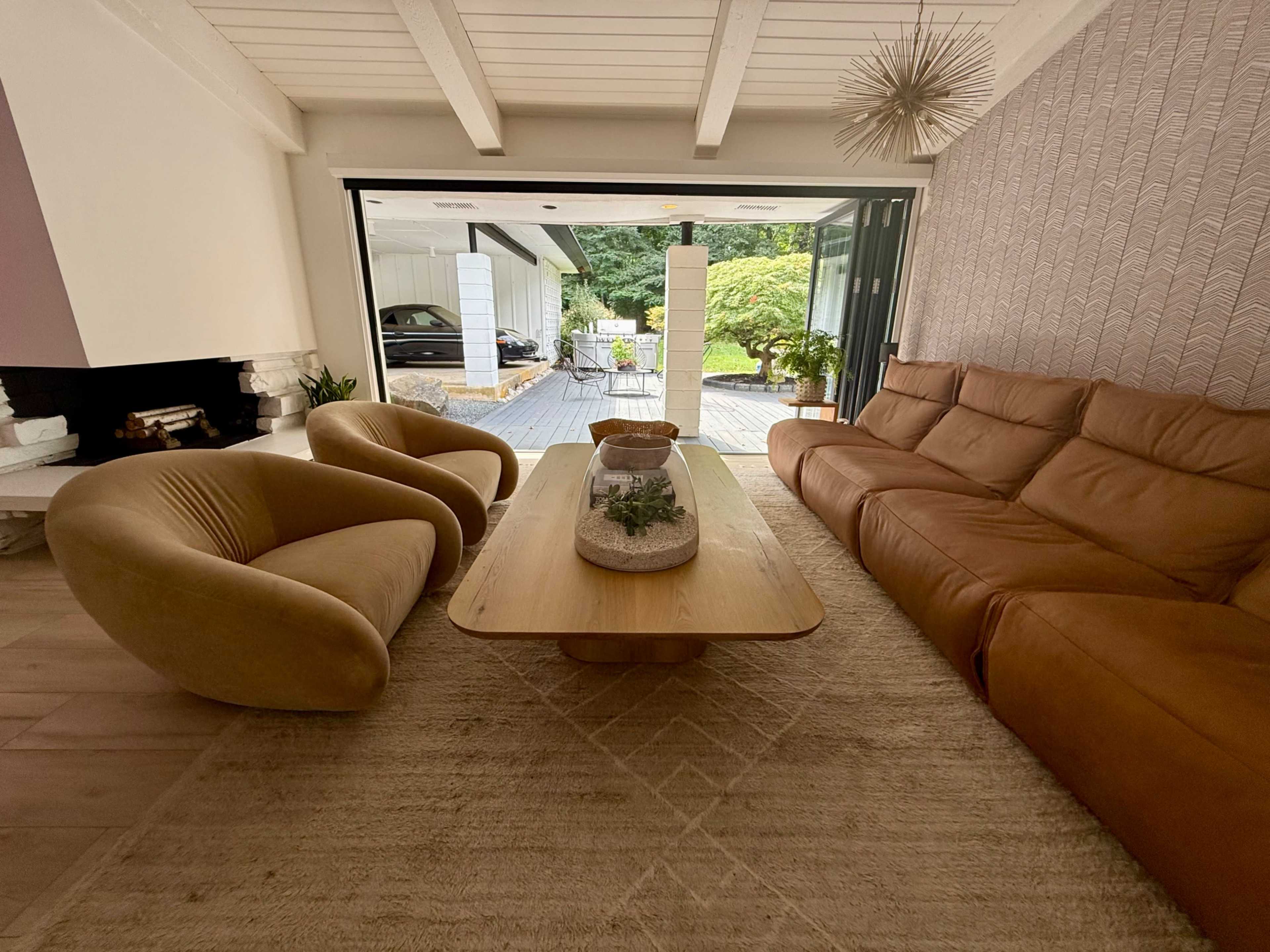The image shows a modern living room with a brown leather sofa, a curved yellow armchair, a wooden coffee table, and a large sliding glass door leading to a patio area.