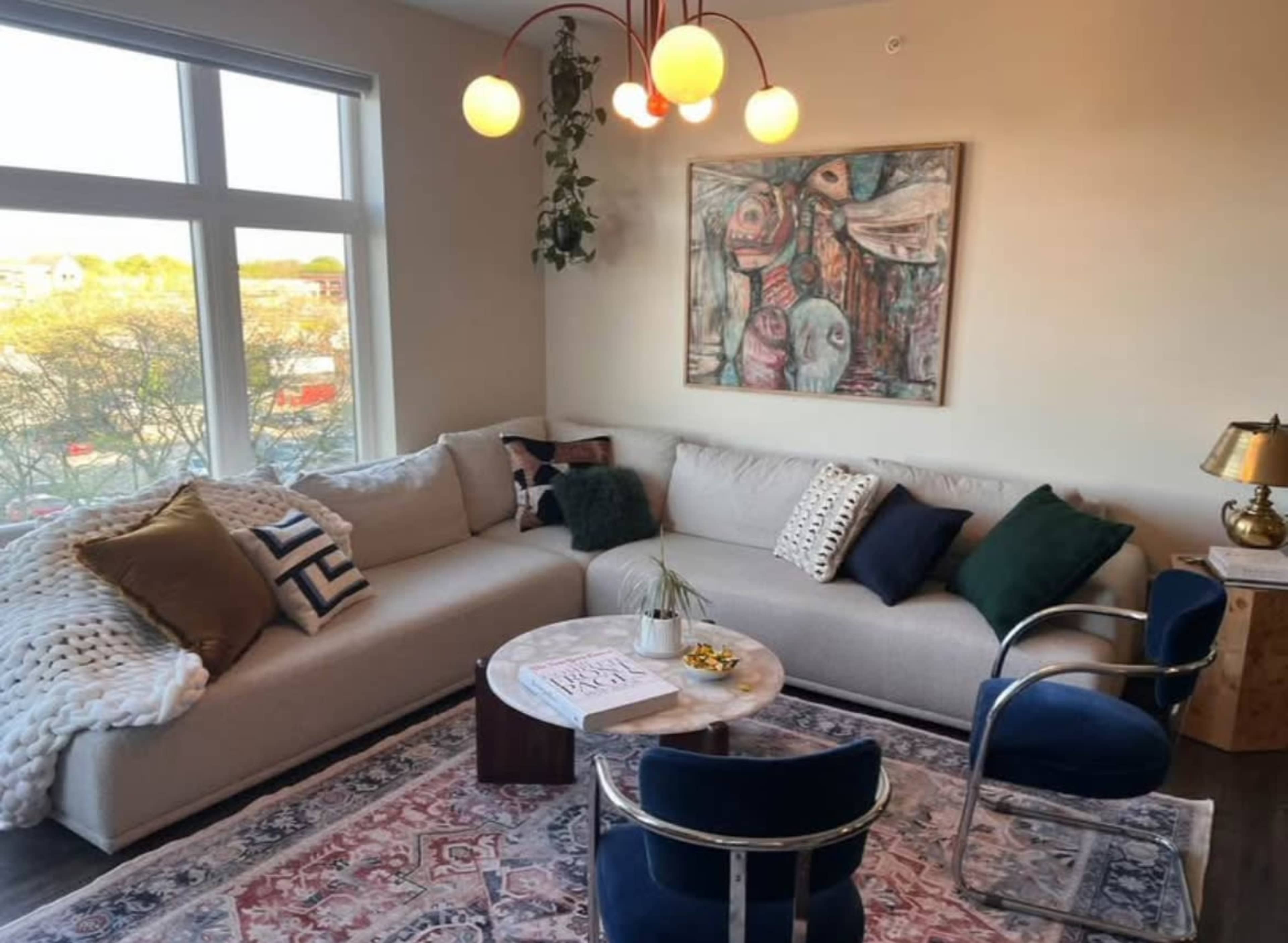 The image shows a cozy living room featuring a light-colored sectional sofa, a round coffee table, a colorful area rug, and wall art, with large windows allowing natural light to fill the space.