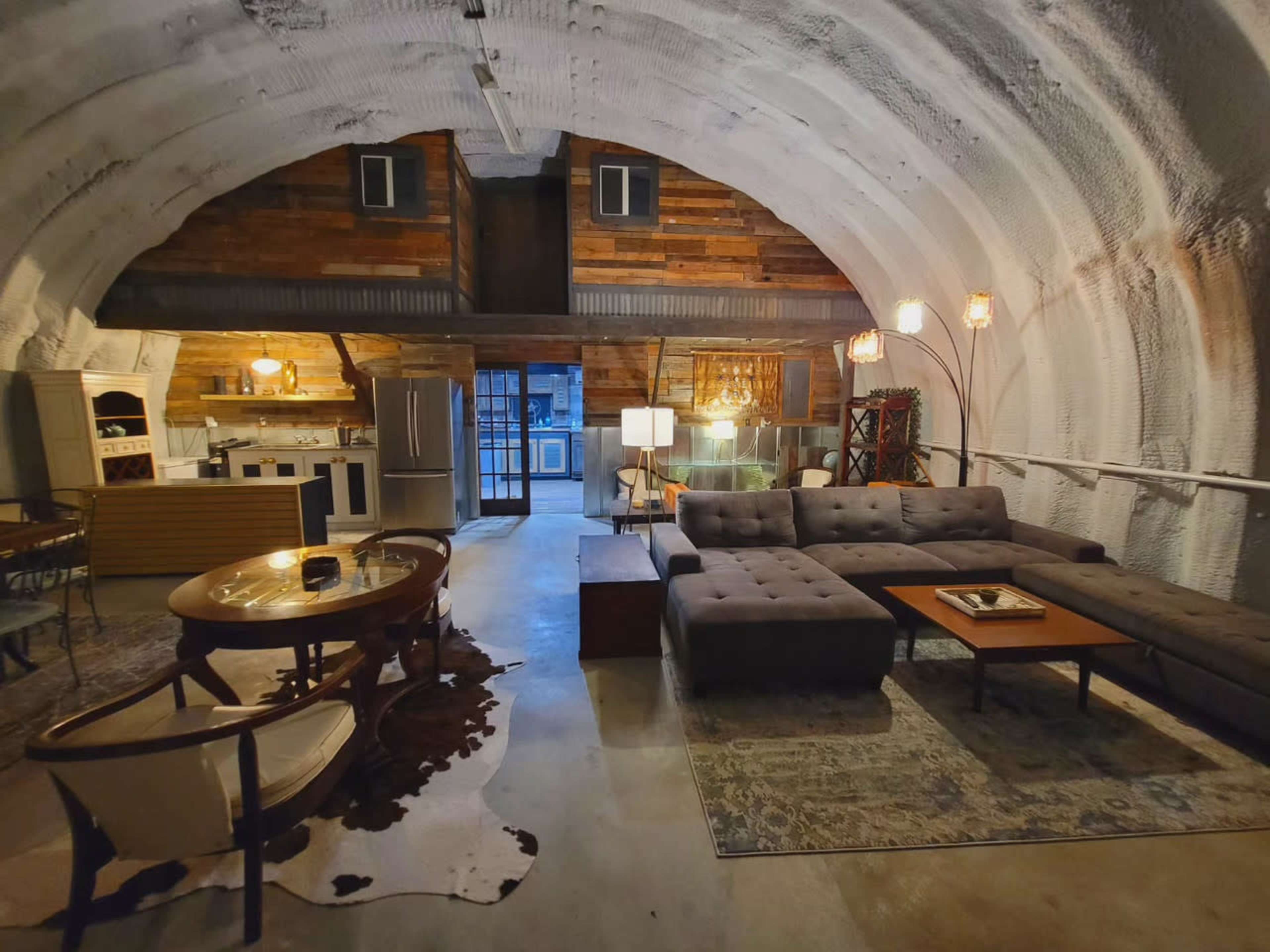 An interior of a modern, arched space featuring a living area with a sectional couch, wooden furniture, and a kitchen in the background.