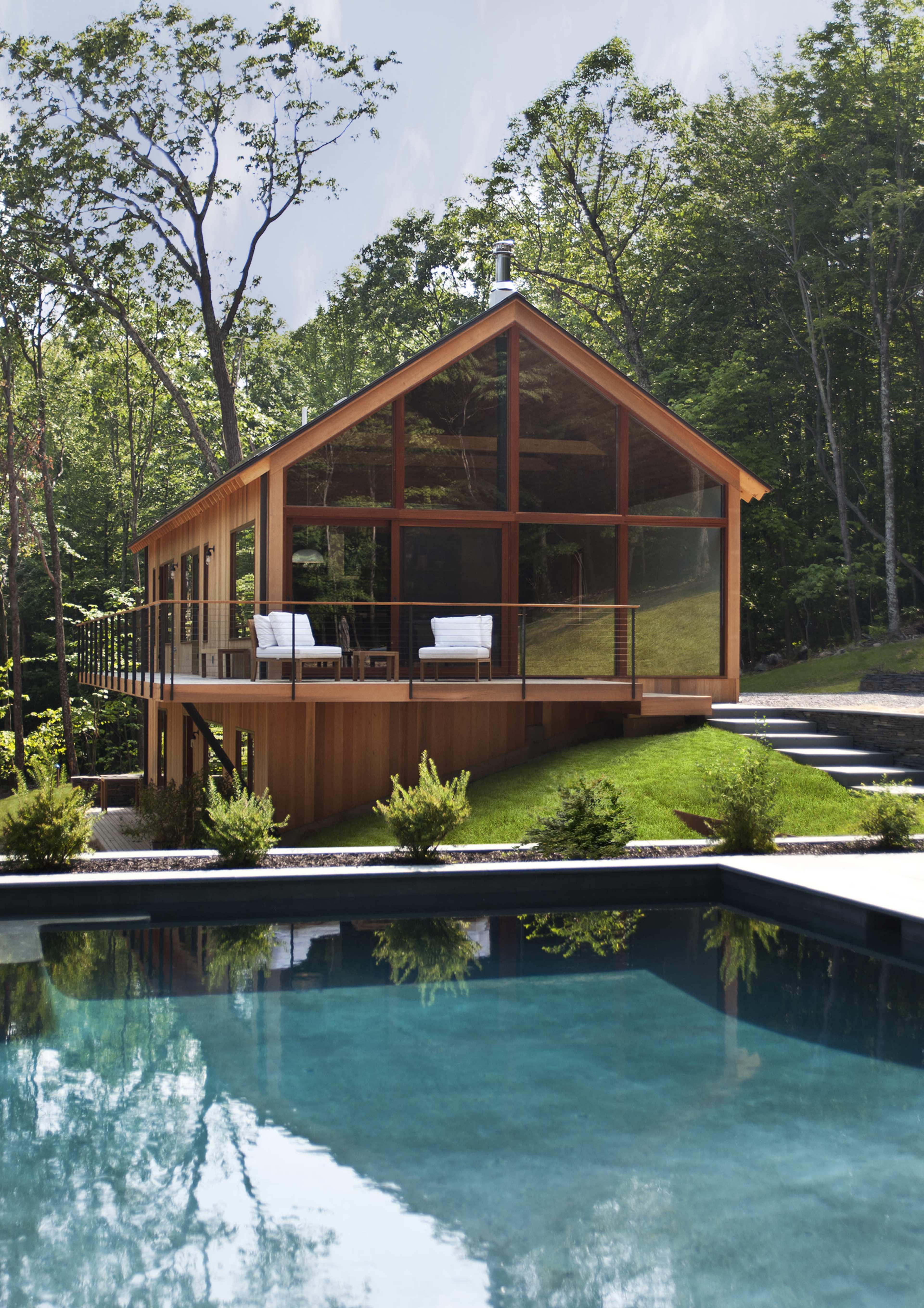 A modern wooden house with large glass windows overlooks a swimming pool surrounded by greenery.